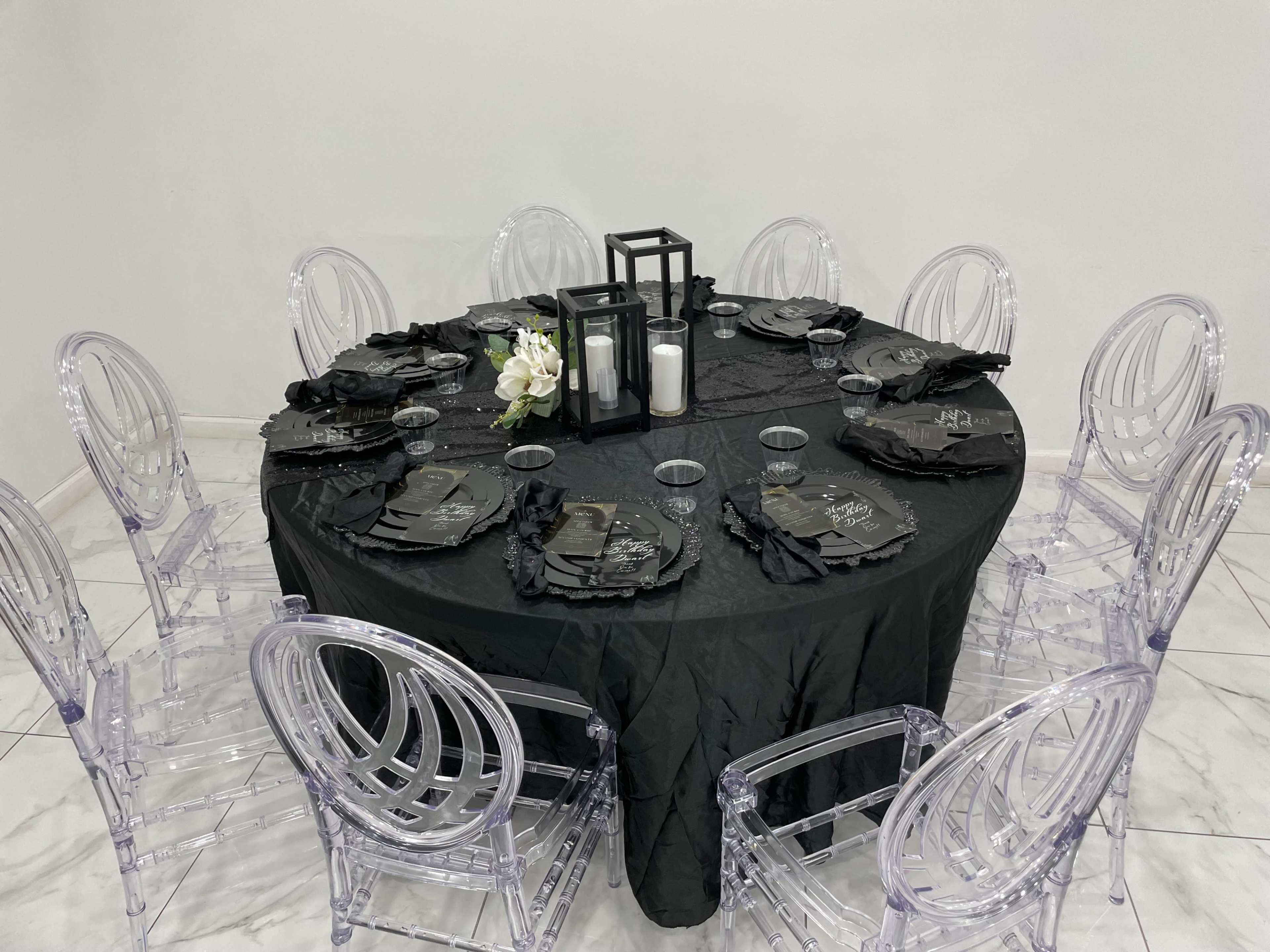 A round table is set with a black tablecloth, clear chairs, black plates, and decorative candle holders, surrounded by twelve chairs.