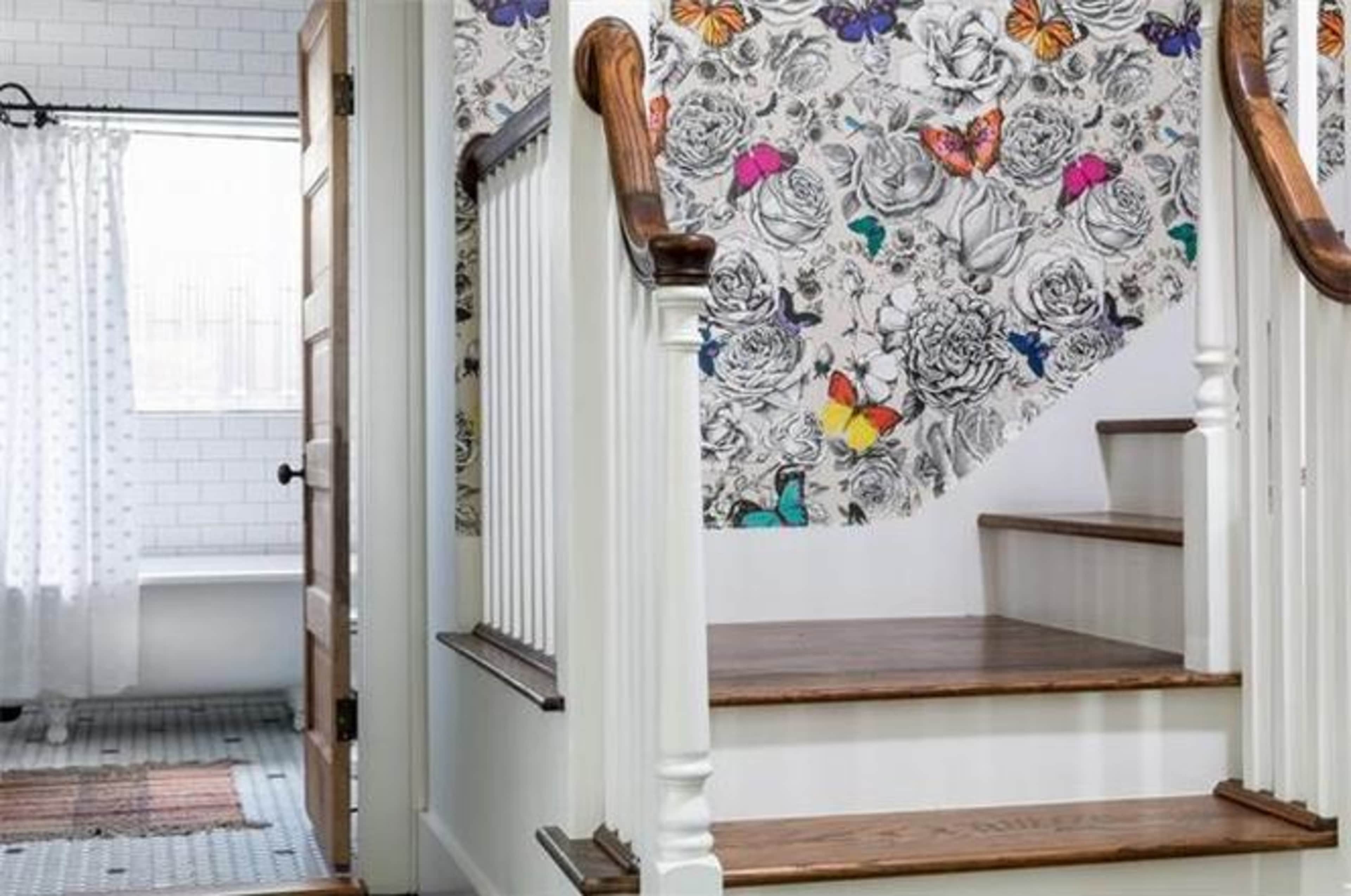 A staircase with a floral and butterfly-patterned wallpaper, leading to a light-filled hallway that connects to a bathroom with a shower curtain.