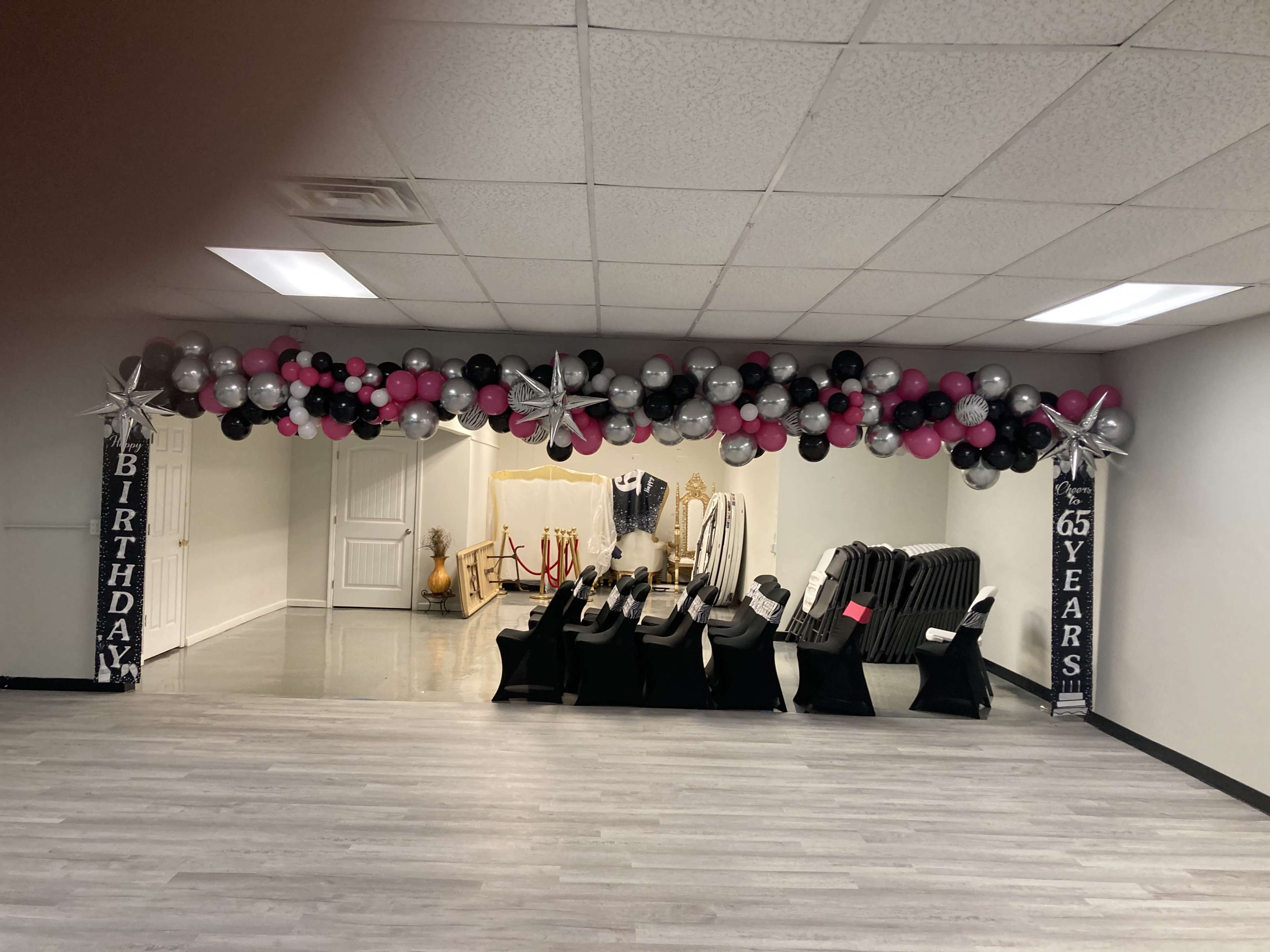 A decorated event space featuring a balloon arch in black, pink, and silver above a row of seating, with signs indicating a 65th birthday celebration.