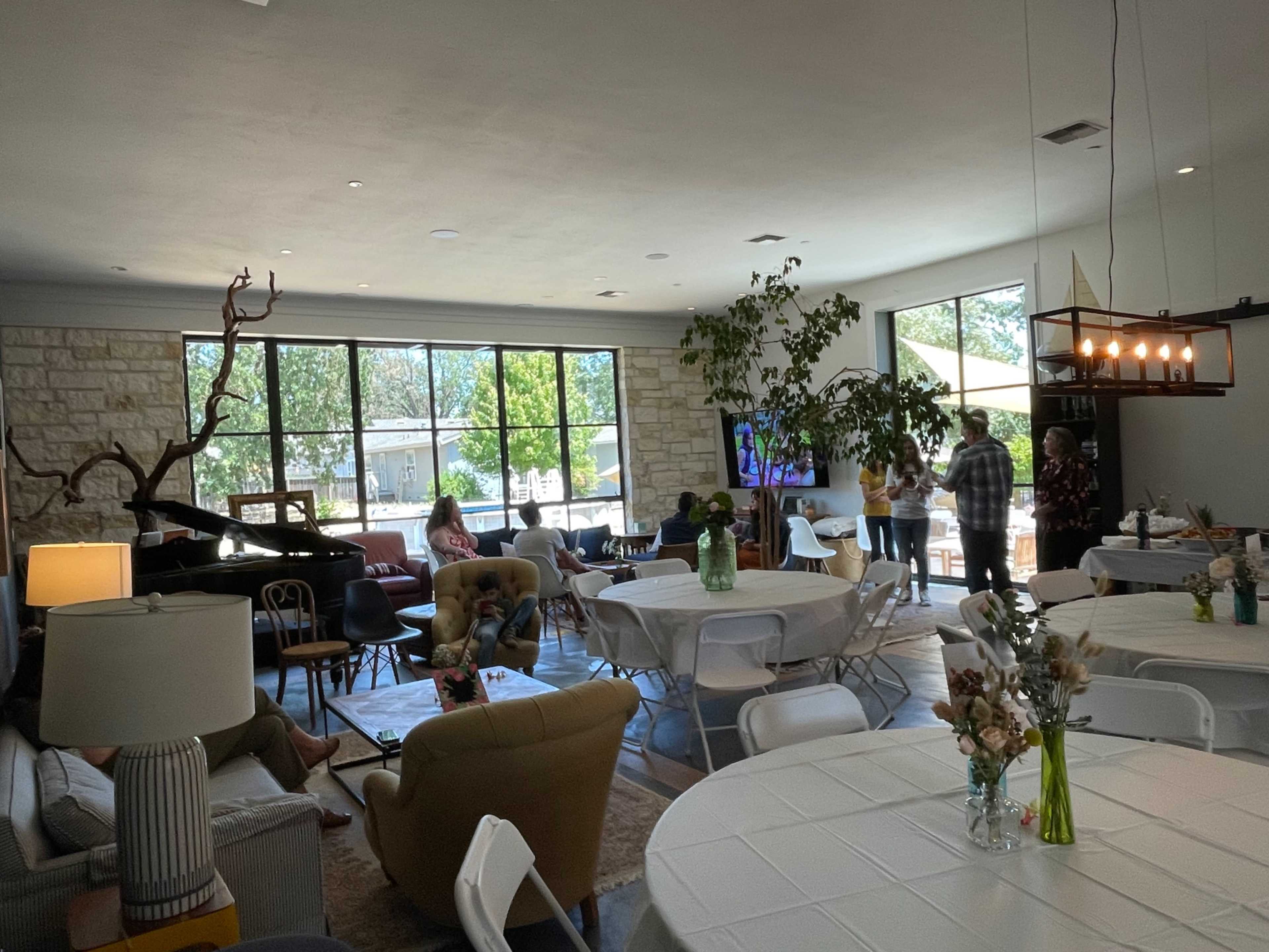 A spacious room with large windows, featuring a mix of seating areas, tables set for gatherings, and a piano in one corner.