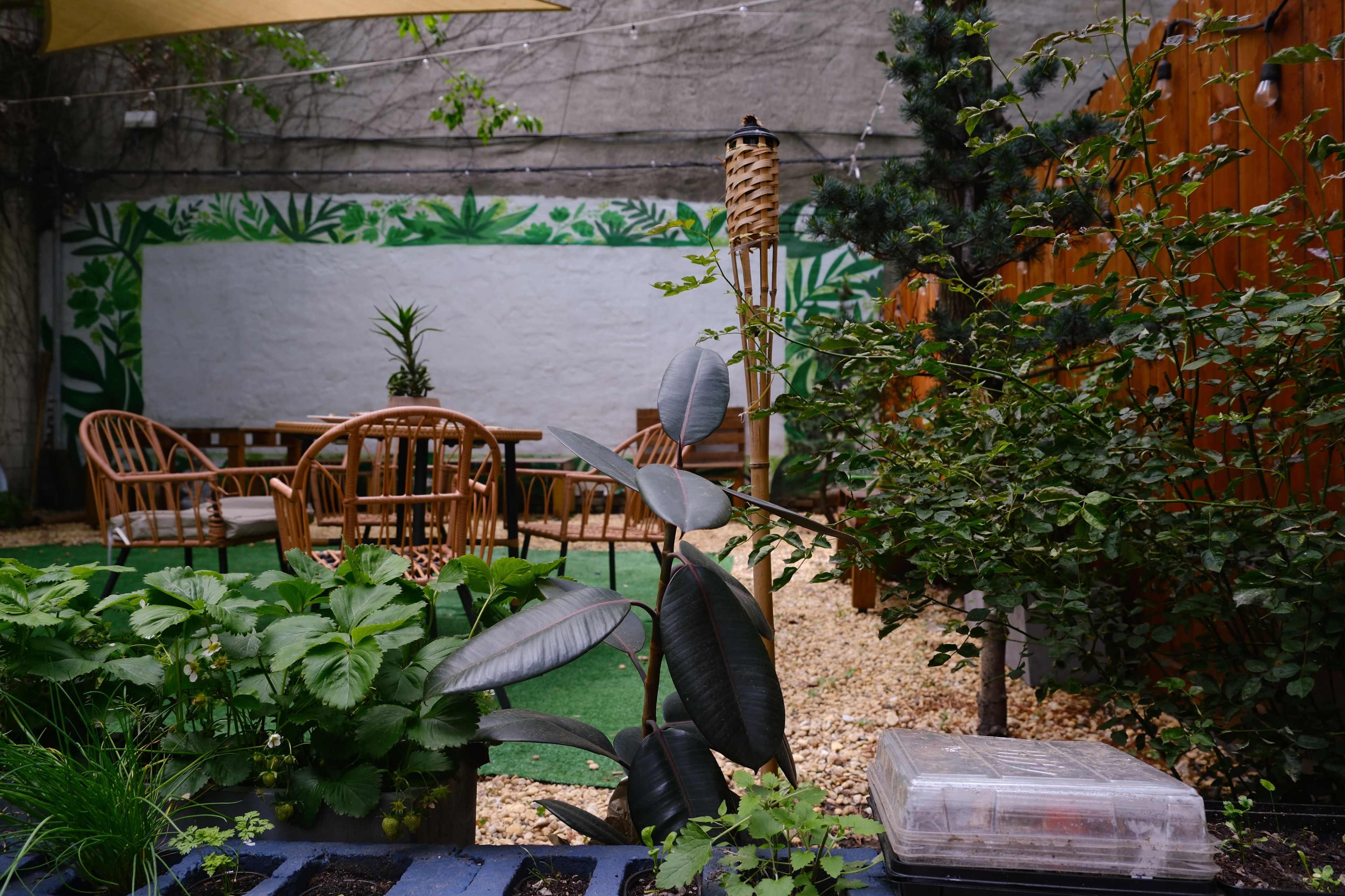 A garden area with wooden chairs and a table surrounded by greenery, including plants and a stone pathway.