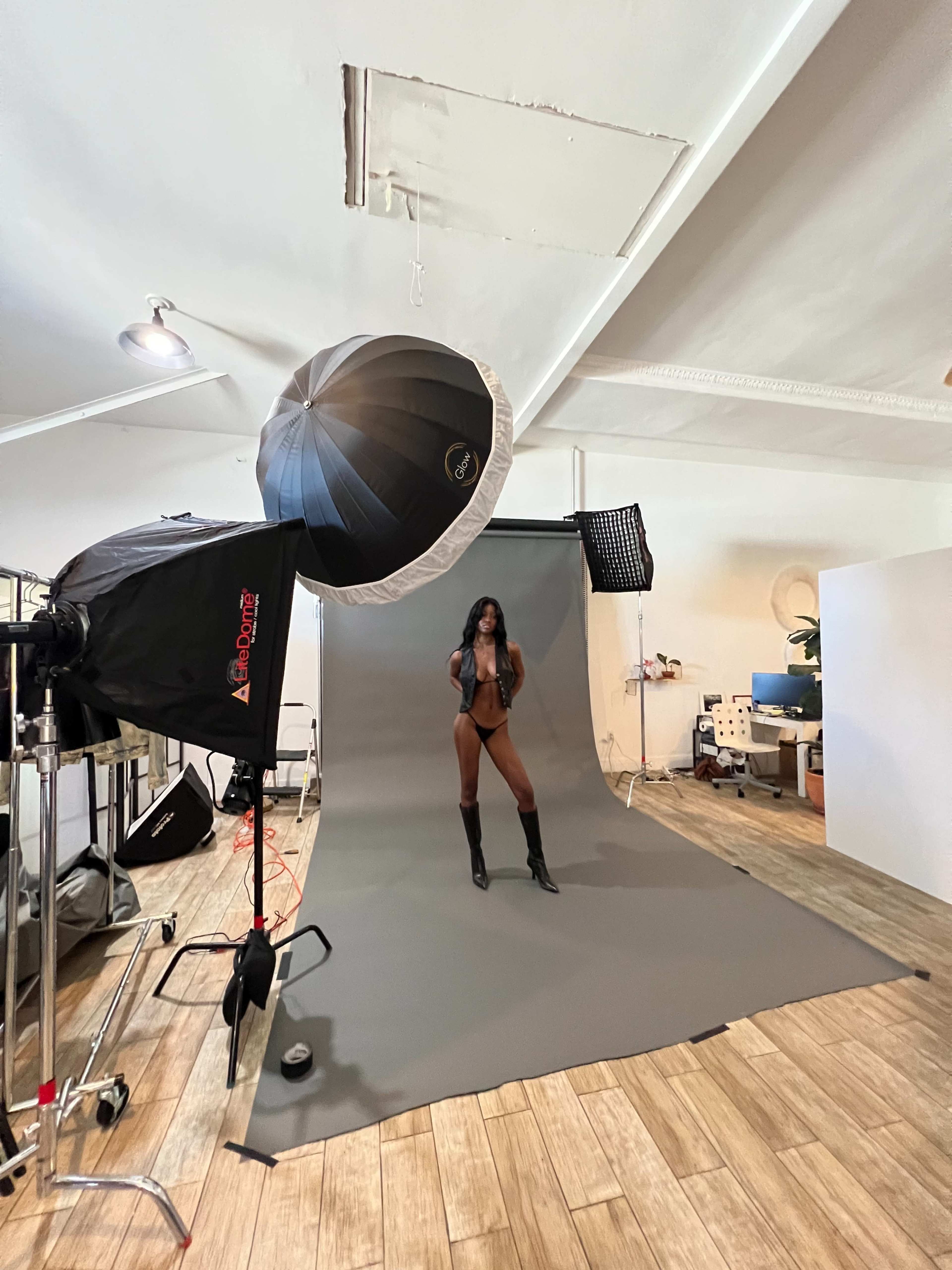 A model poses on a gray backdrop in a photography studio equipped with lighting and camera equipment.