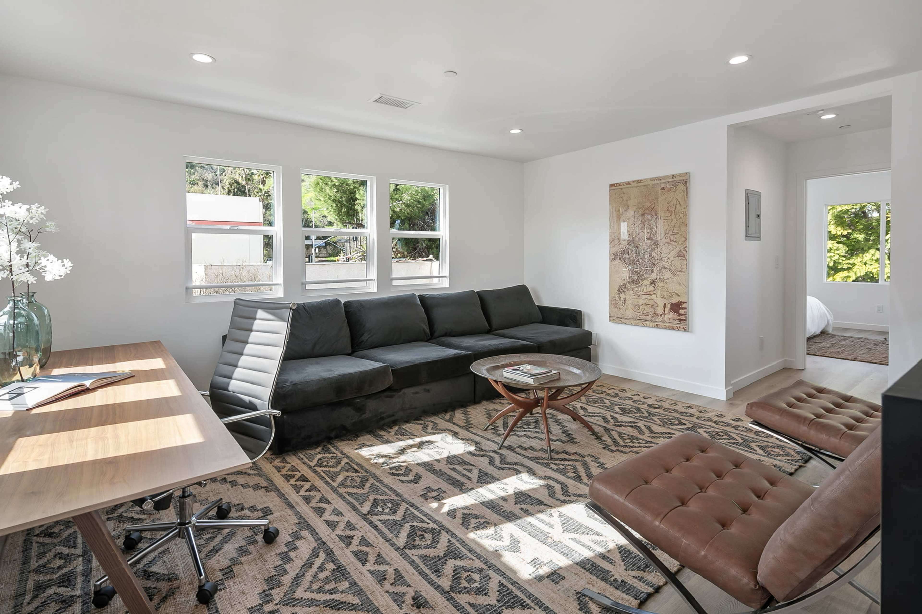 A modern living area with a dark sectional sofa, a glass coffee table, a wooden desk with an office chair, and two leather lounge chairs, all set against a neutral-colored wall.
