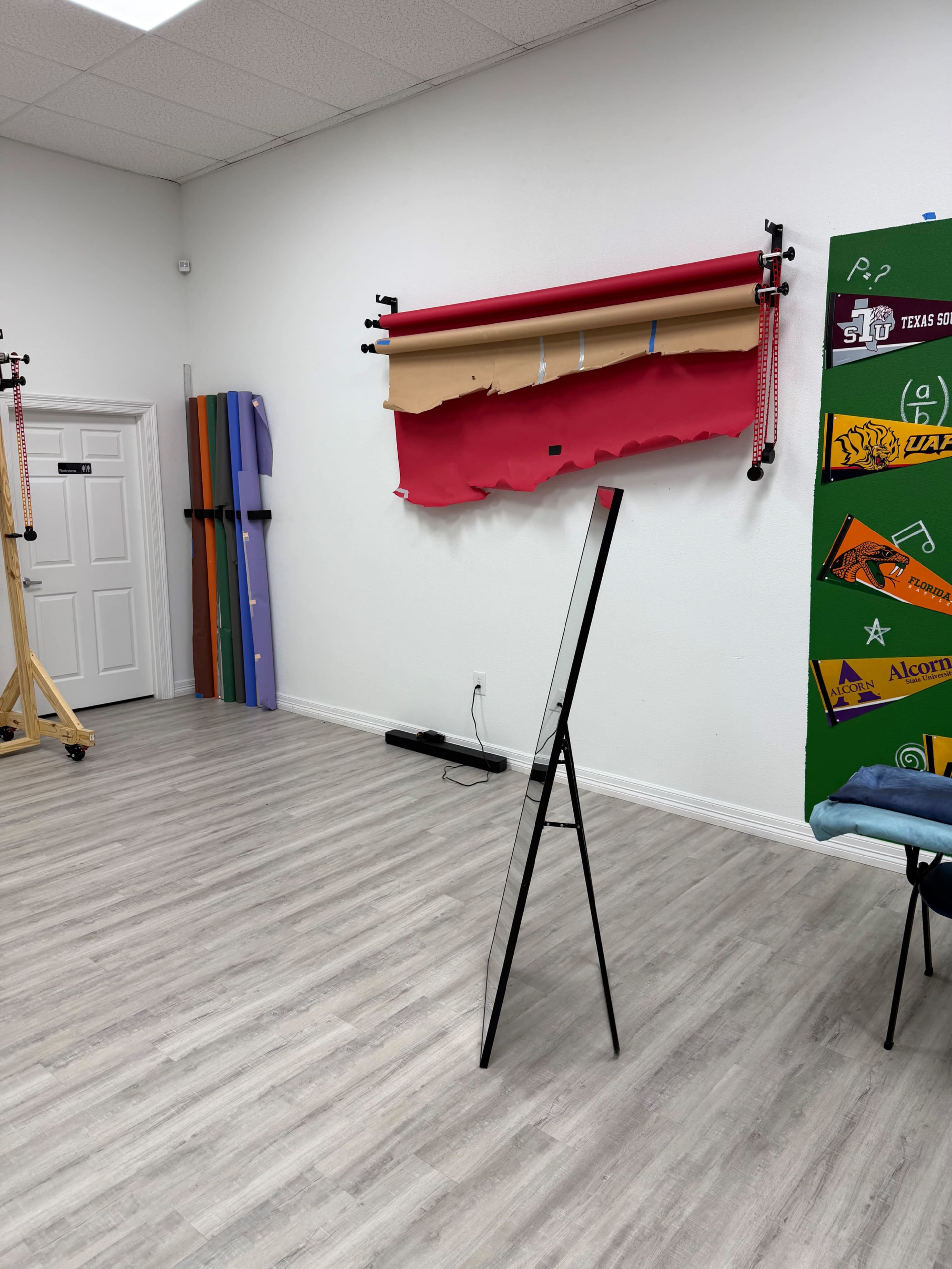 The image shows a bright, empty studio space with a wooden easel, colorful backdrops hanging on the wall, and a green display board featuring various items.
