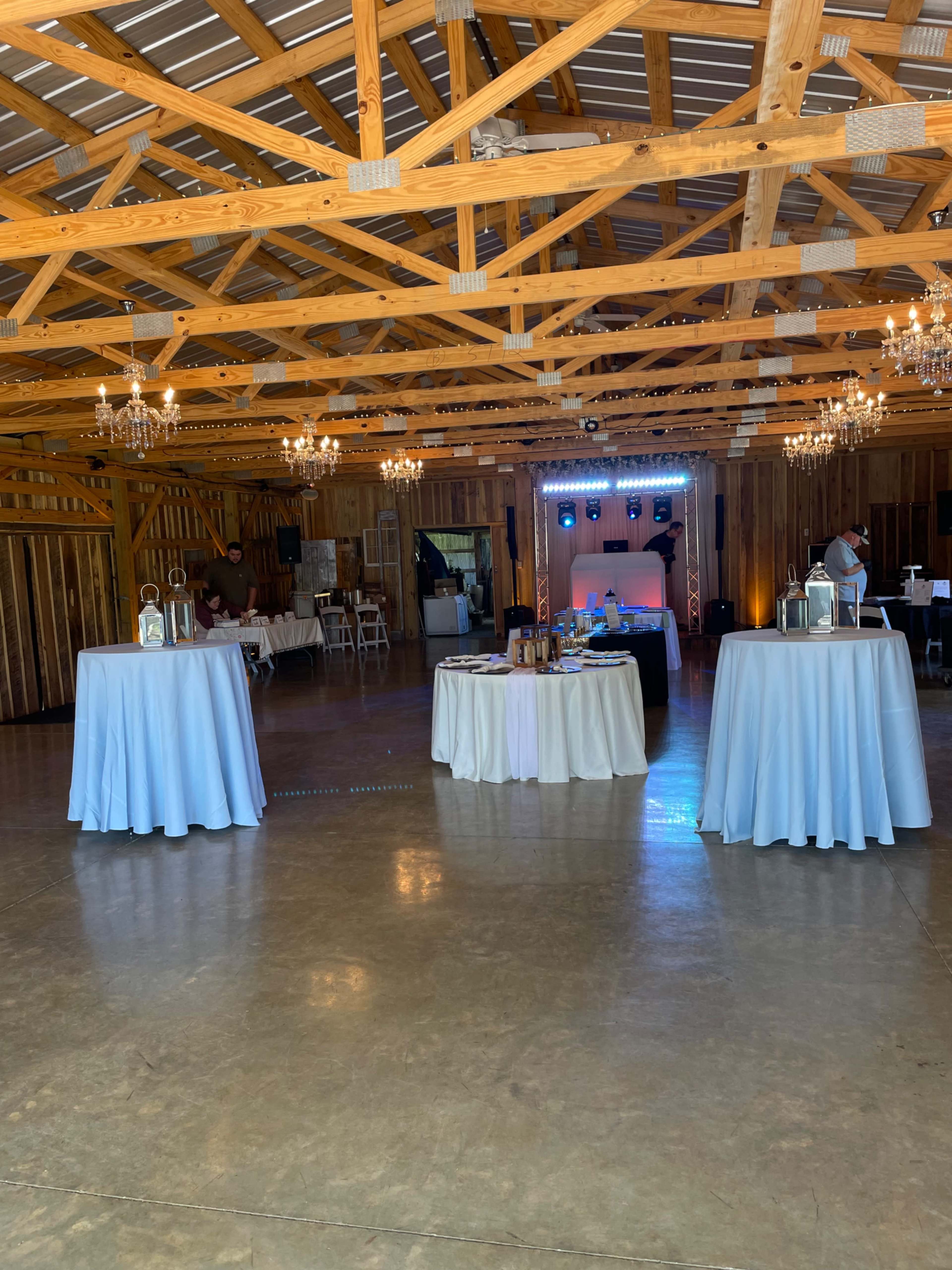 The interior of a barn-style venue features wooden beams, chandeliers, and several tables dressed in light blue linens, arranged in an open space.