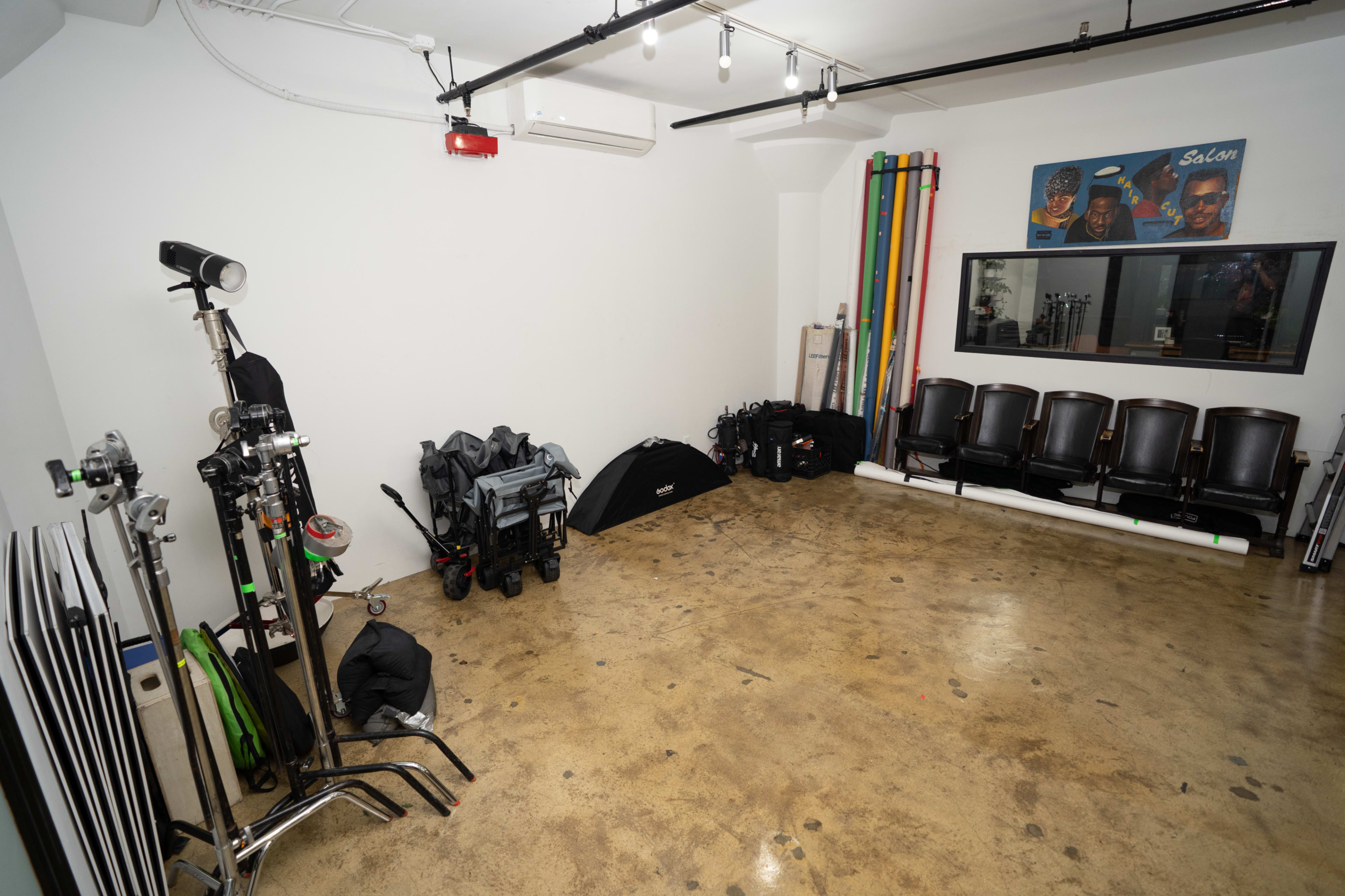 The image shows an empty studio space with various photography equipment and black chairs arranged against the wall.