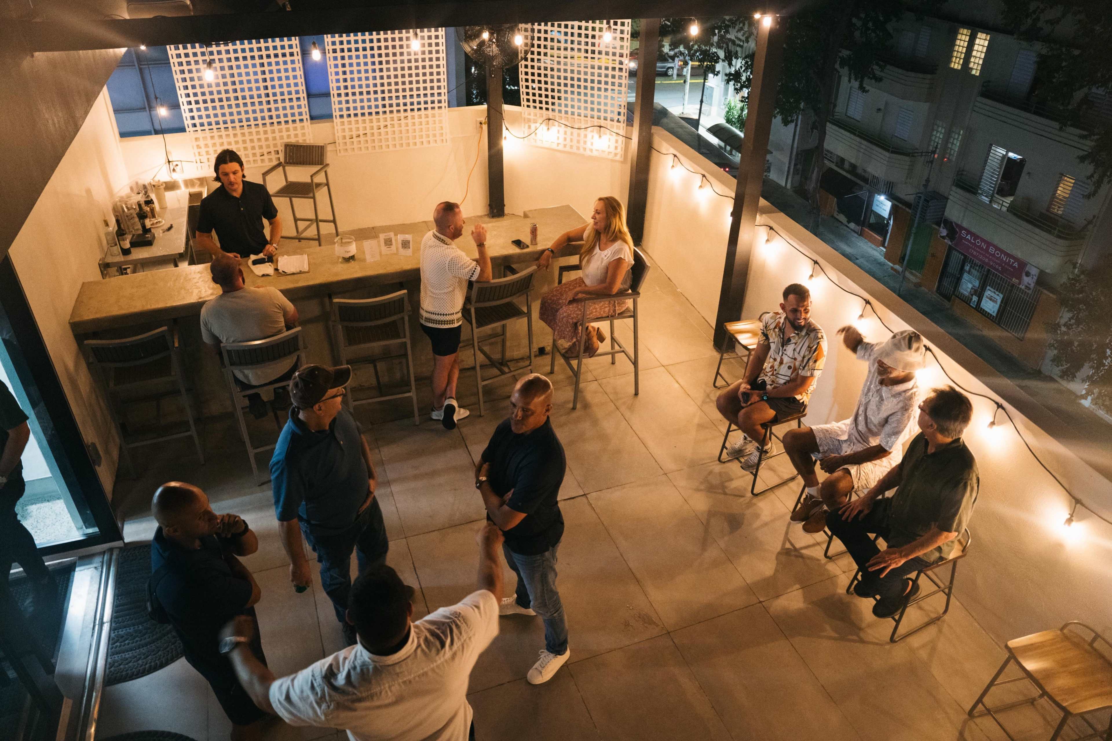 A rooftop bar scene features a mix of seated and standing patrons enjoying drinks, with a bartender in the background under string lights.