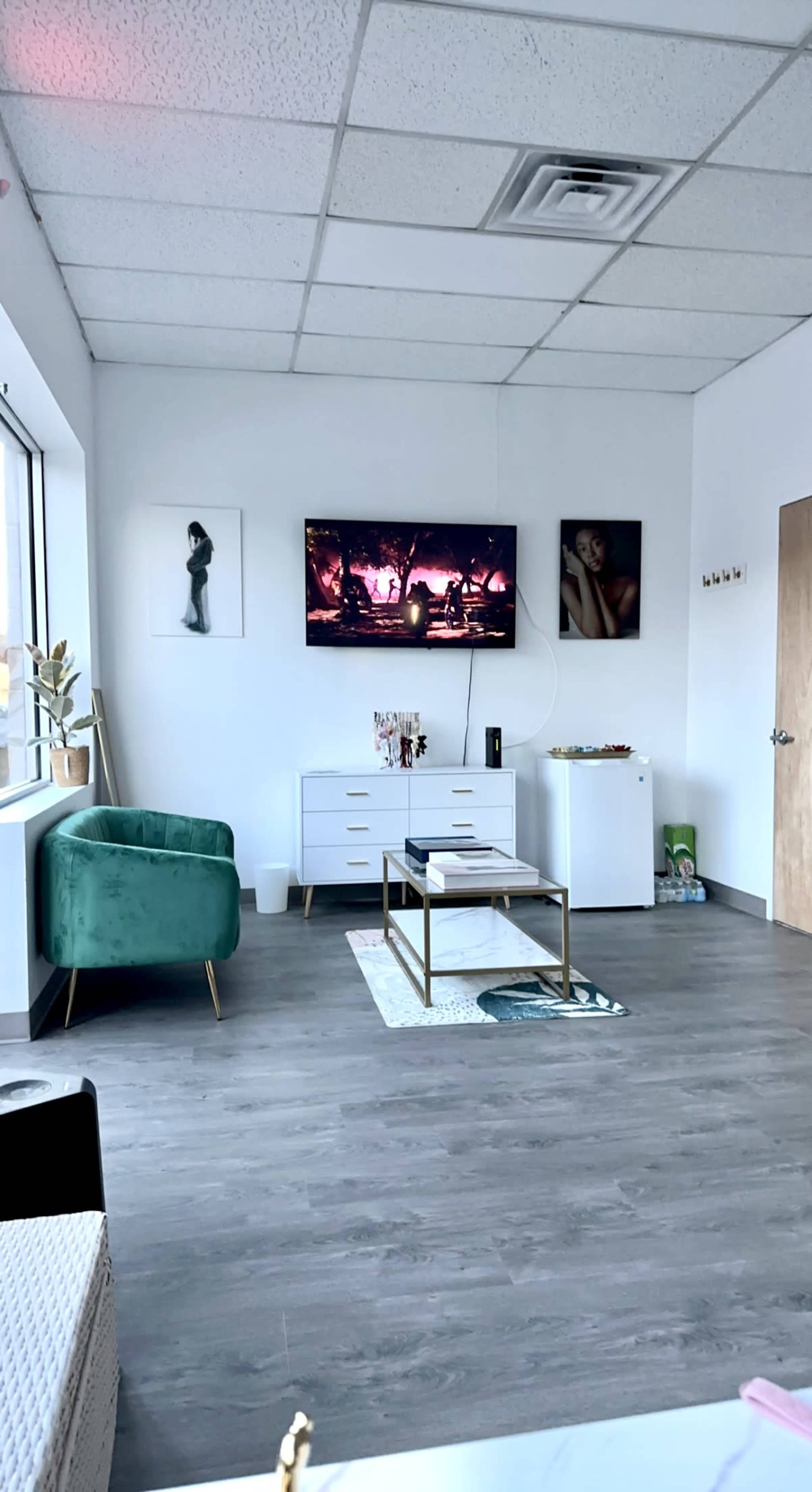A modern, minimalist room featuring a green chair, a white dresser, and a television mounted on the wall, with framed artwork and a small table in the center.