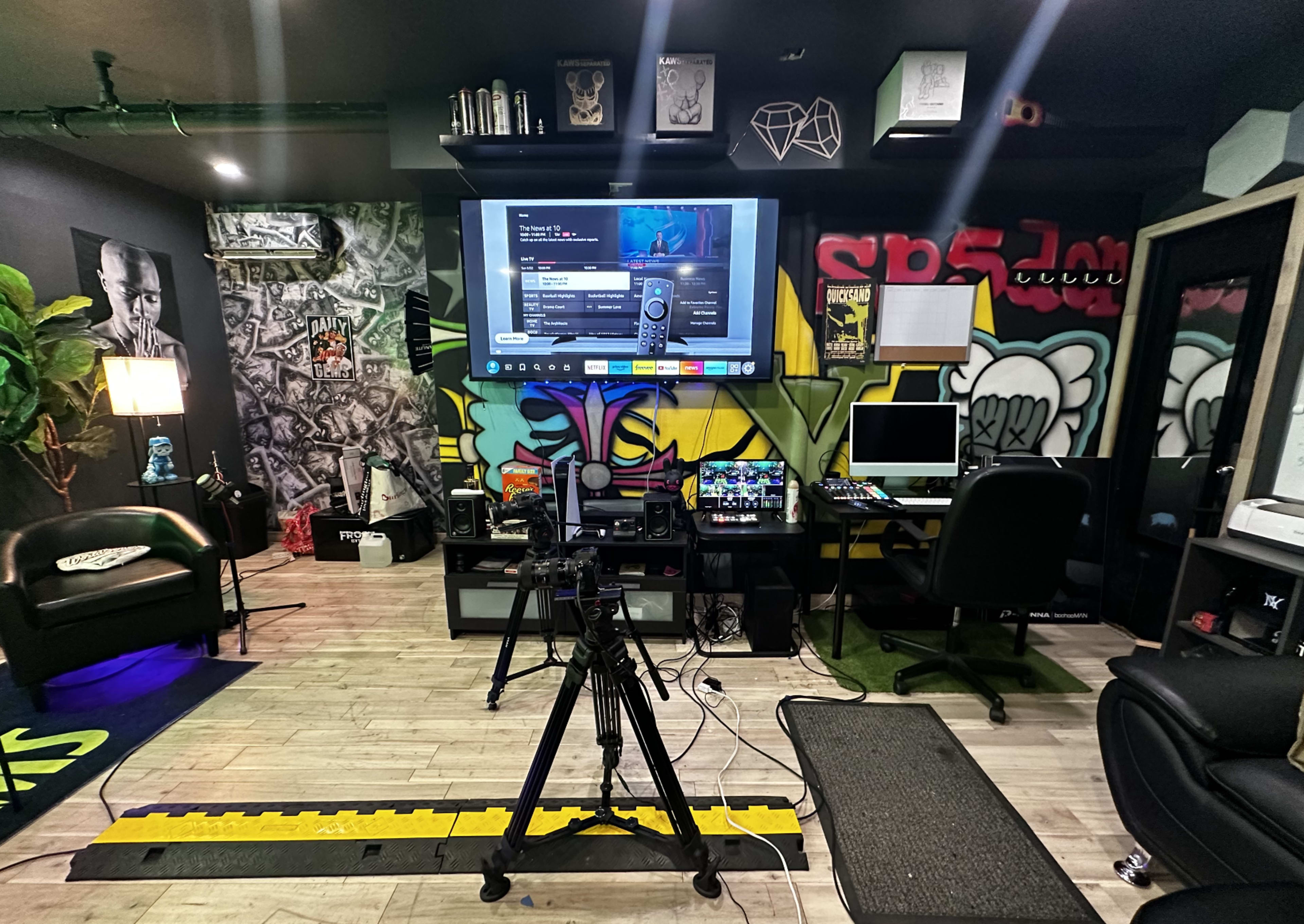 A multimedia production room with a large screen displaying a video interface, a camera on a tripod, and various equipment and furniture arranged against colorful graffiti-style walls.