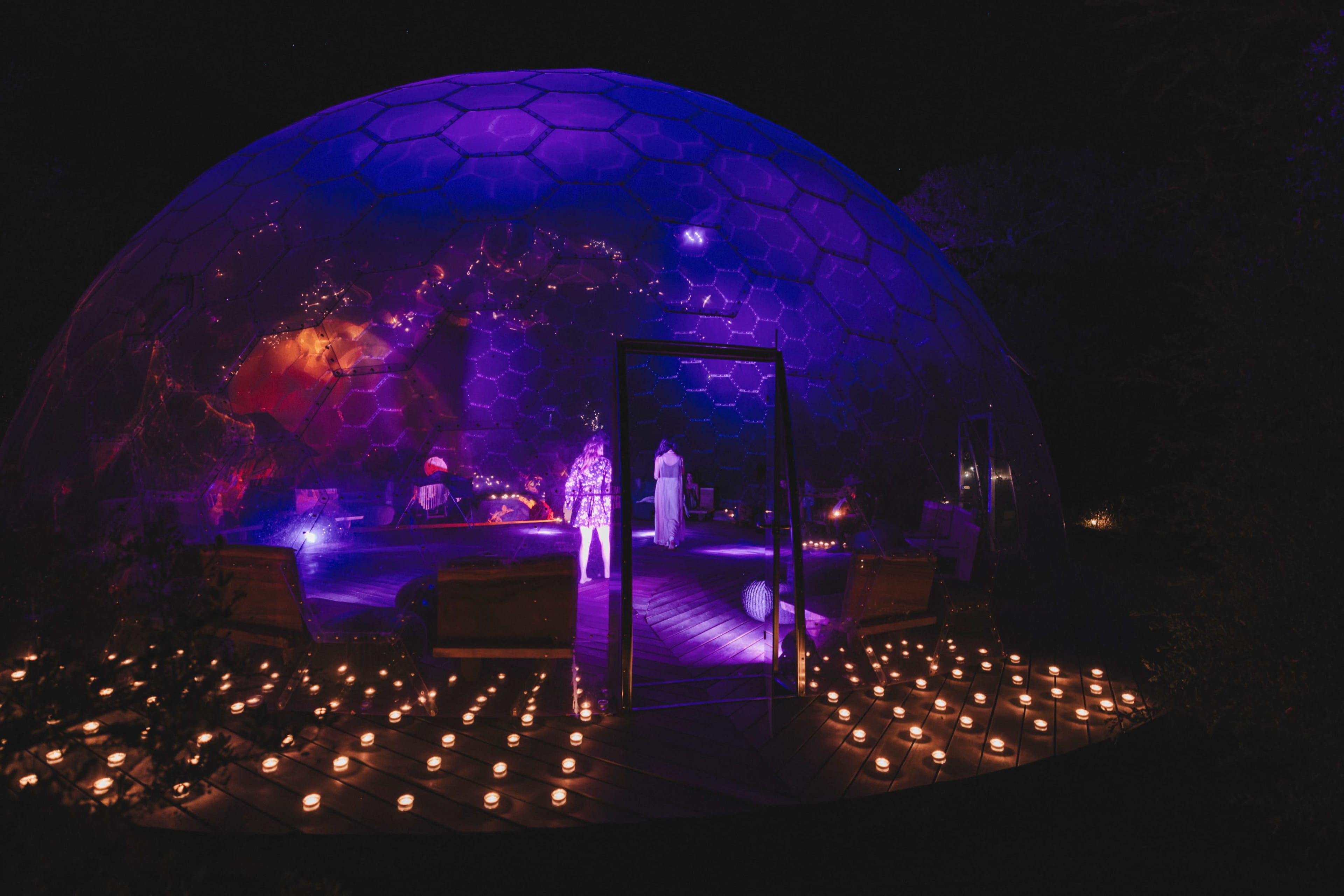 A geodesic dome illuminated by purple lights is surrounded by candles on a wooden deck, set against a night sky.