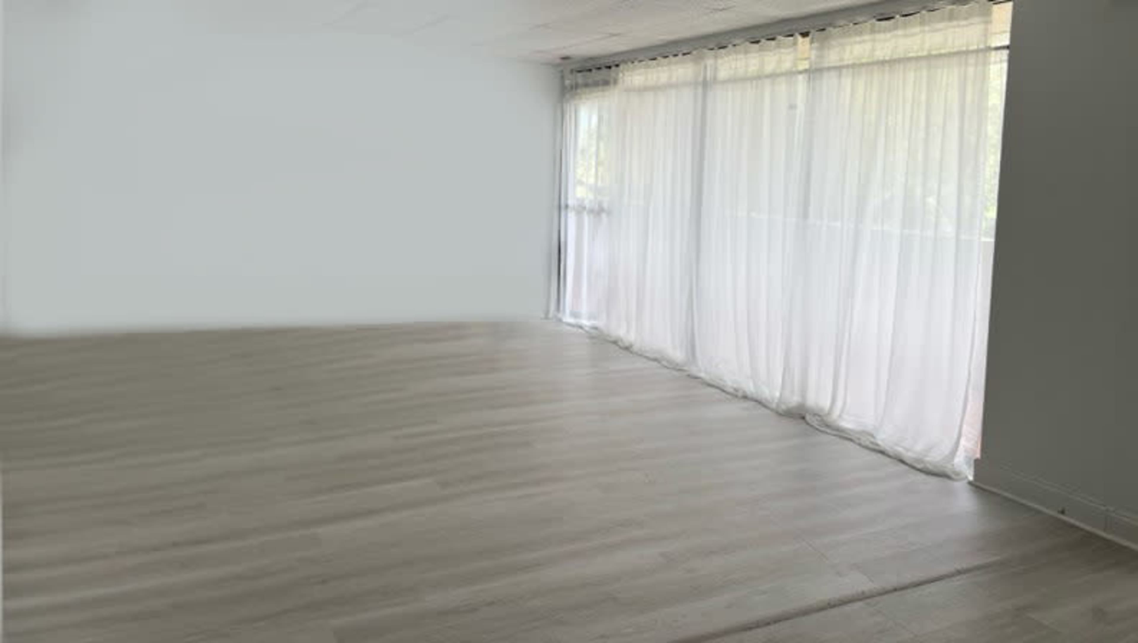 The image shows a spacious room with light wooden flooring and sheer curtains covering a large window.