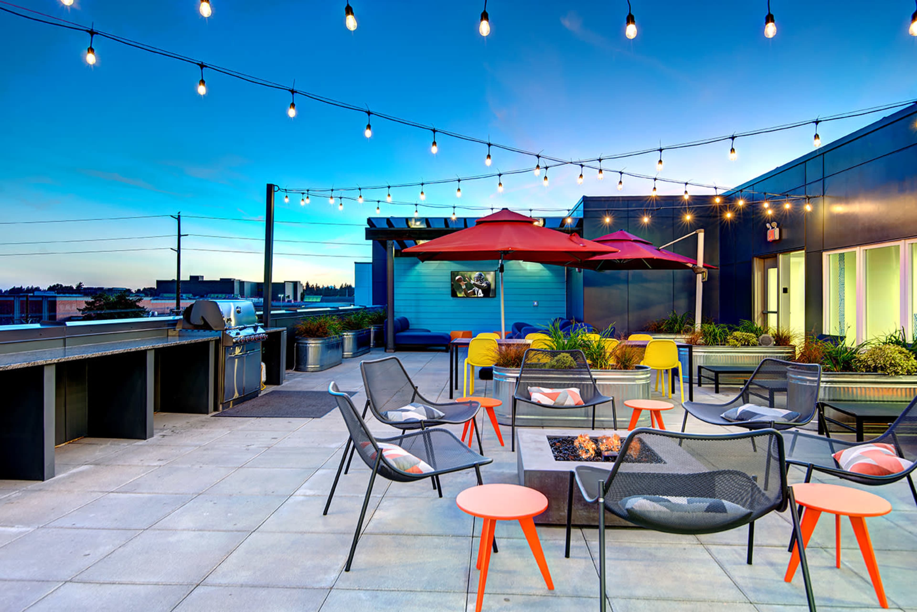 A rooftop terrace adorned with string lights, featuring seating arrangements around a fire pit, tables, and patio umbrellas.