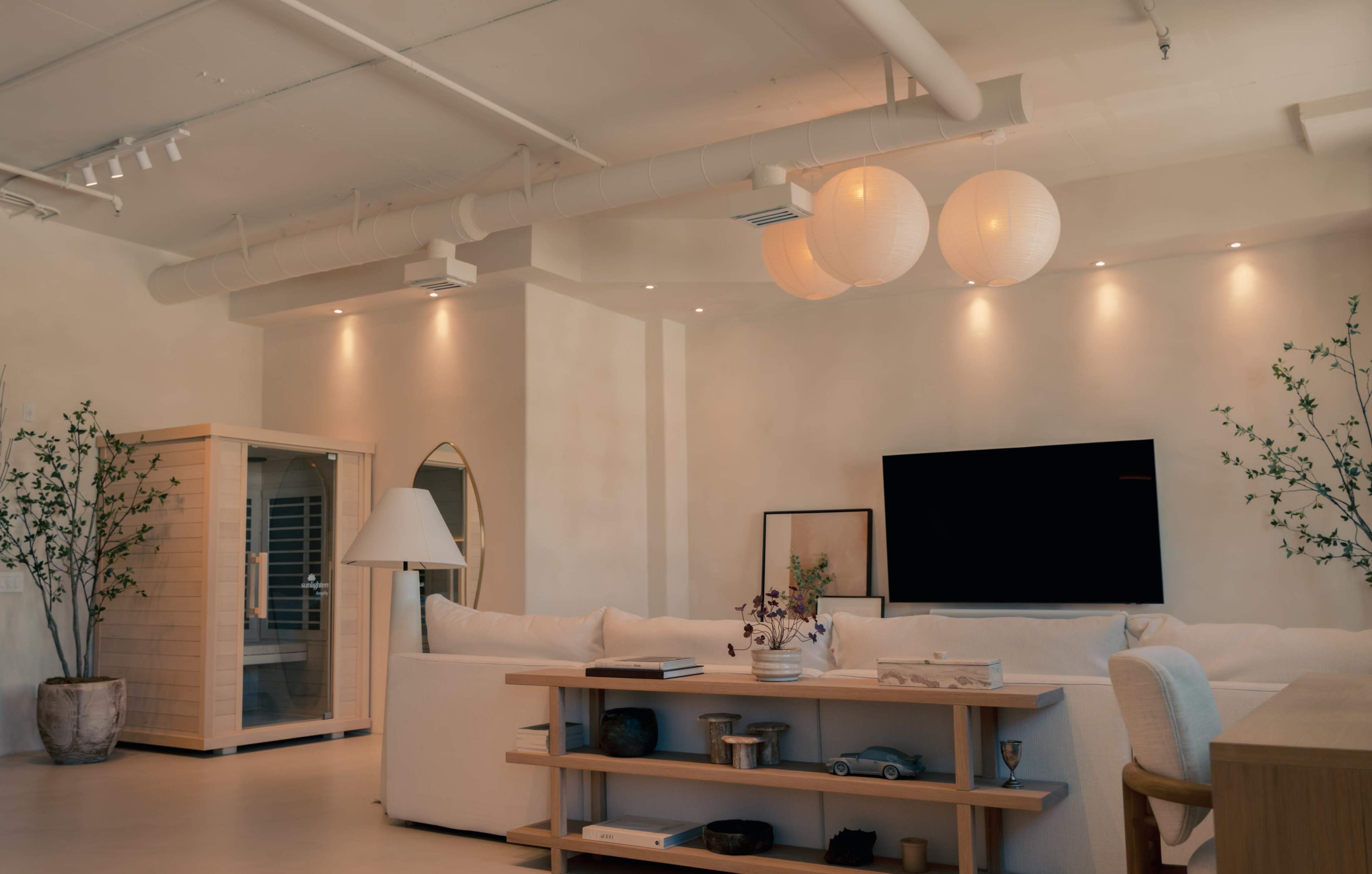 The image shows a modern living room featuring a sectional sofa, a wall-mounted television, ambient lighting, and a sauna in the corner.