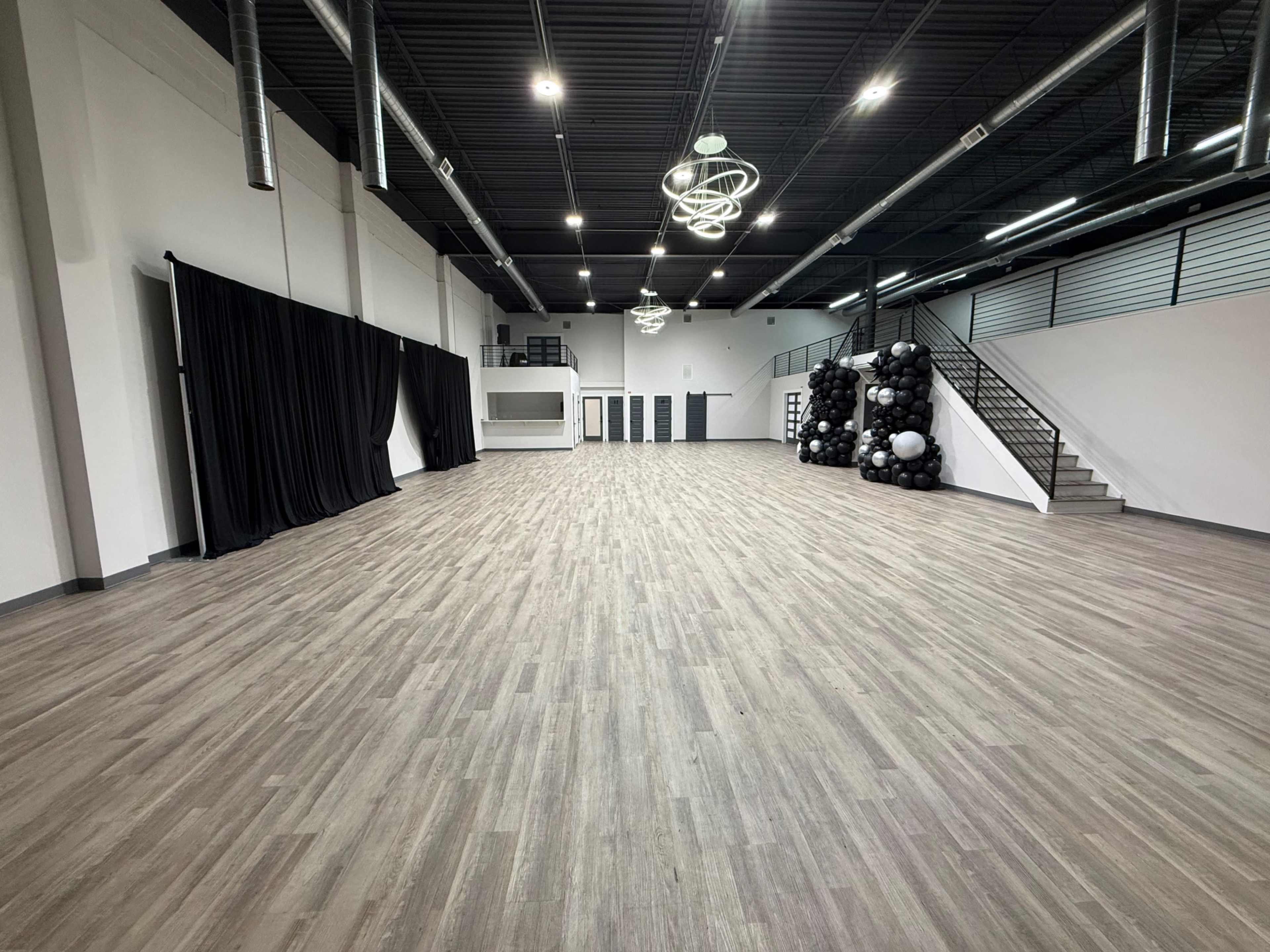 The image shows a spacious, empty interior room with wooden flooring, large windows, and a staircase, featuring black curtains on one side and a cluster of black balloons arranged on the other.