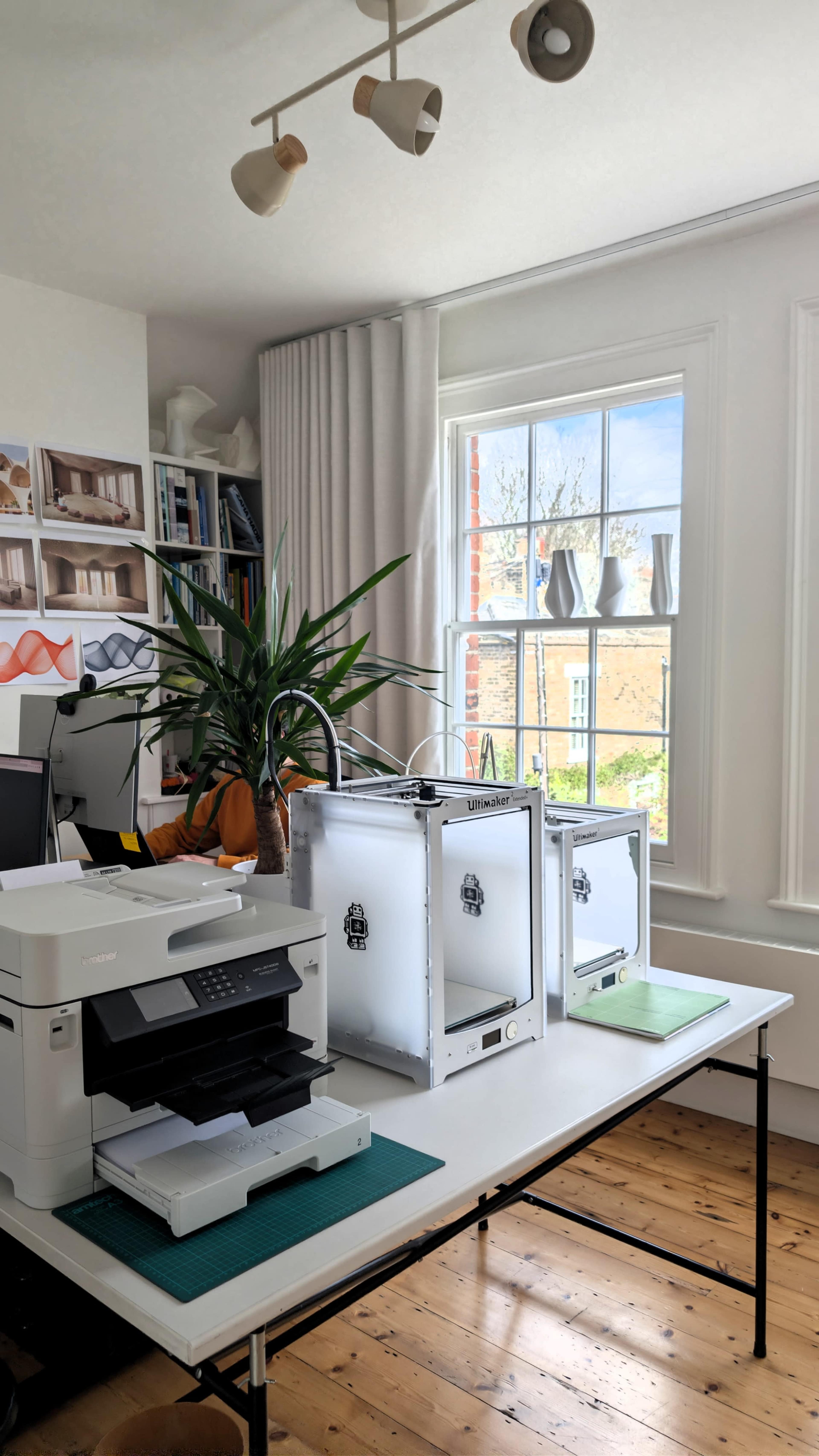 The image shows a bright office space with a printer, three 3D printers, and a large window that overlooks an outdoor area.