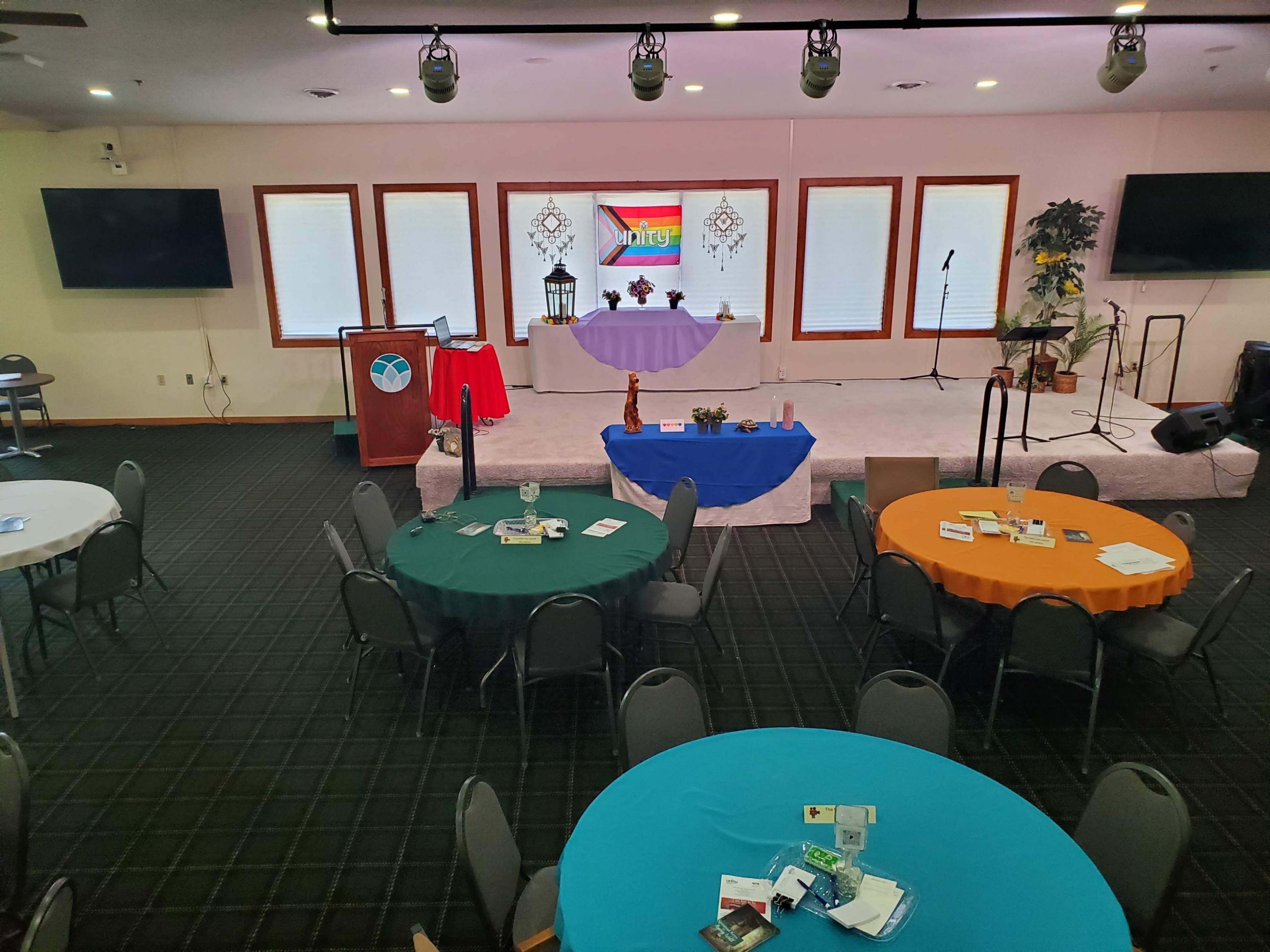 A spacious indoor event venue with several round tables covered in colorful tablecloths, a stage at the front with a podium, and decorations set for a gathering.
