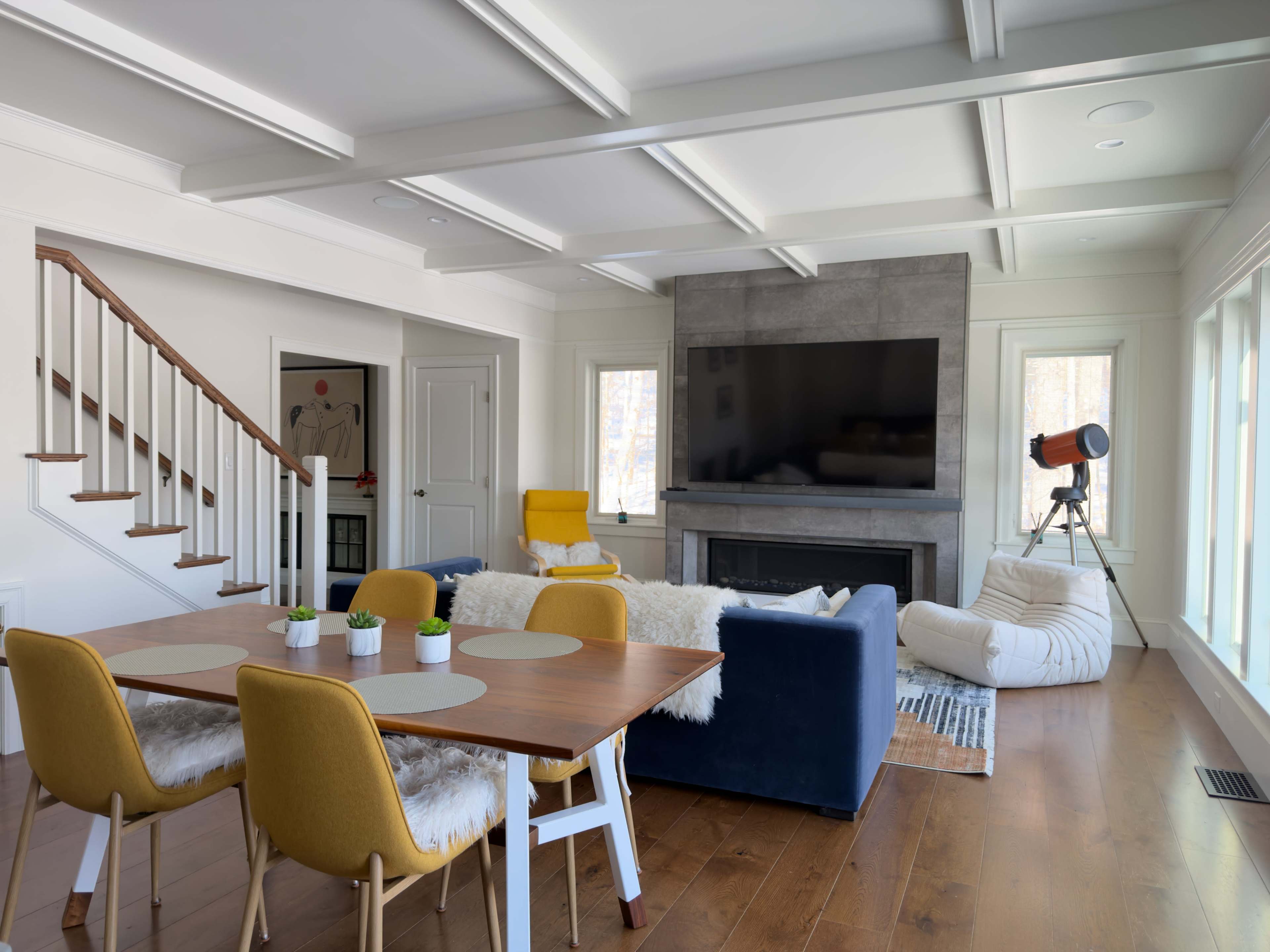 The image shows a modern living space featuring a dining area with a wooden table and yellow chairs, a cozy seating area with a blue couch and decorative throw, and a staircase leading to the upper level, accompanied by large windows and a mounted television.