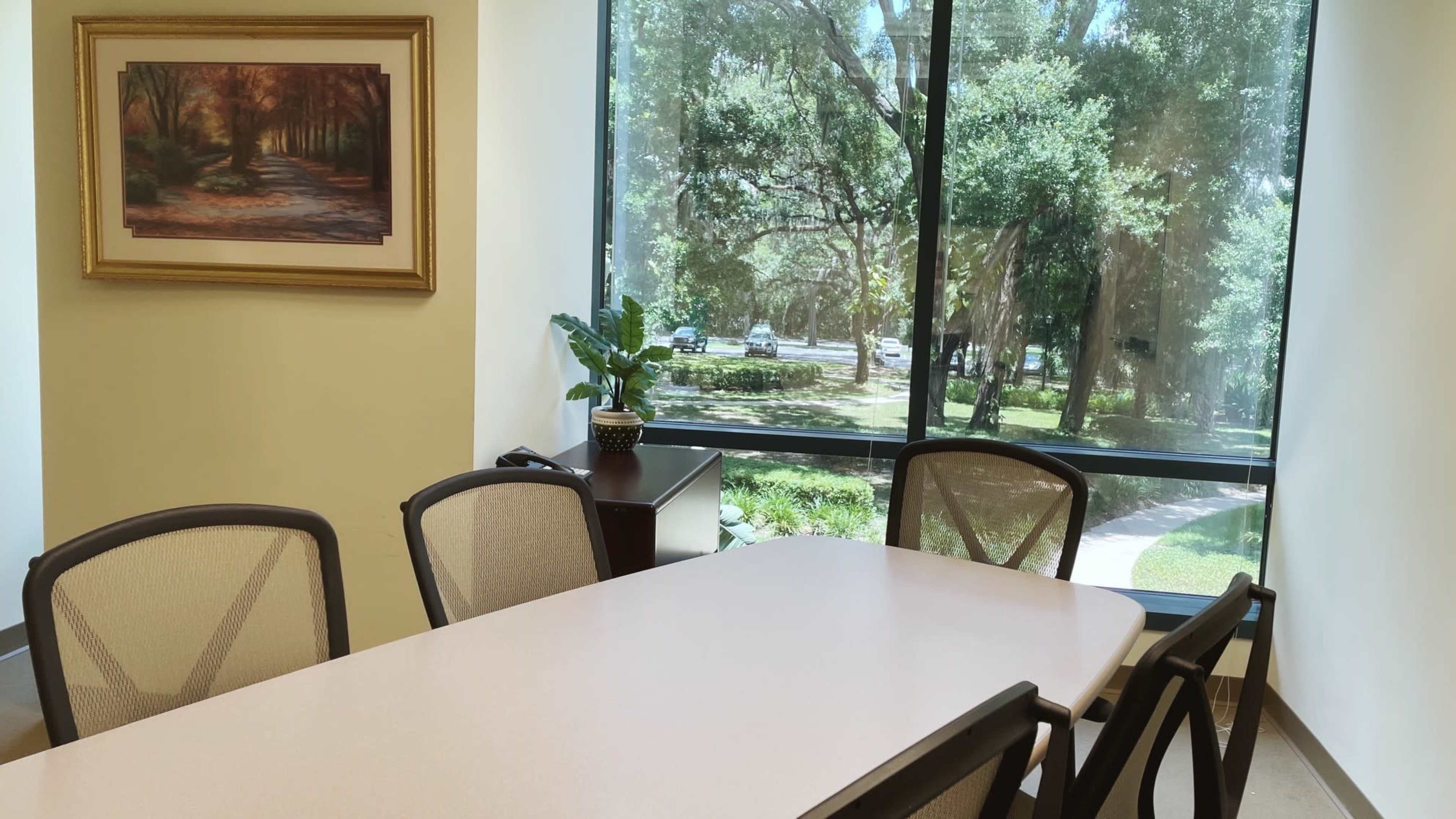 A conference room with a large table and chairs, featuring a view of trees and a landscaped area through a large window.