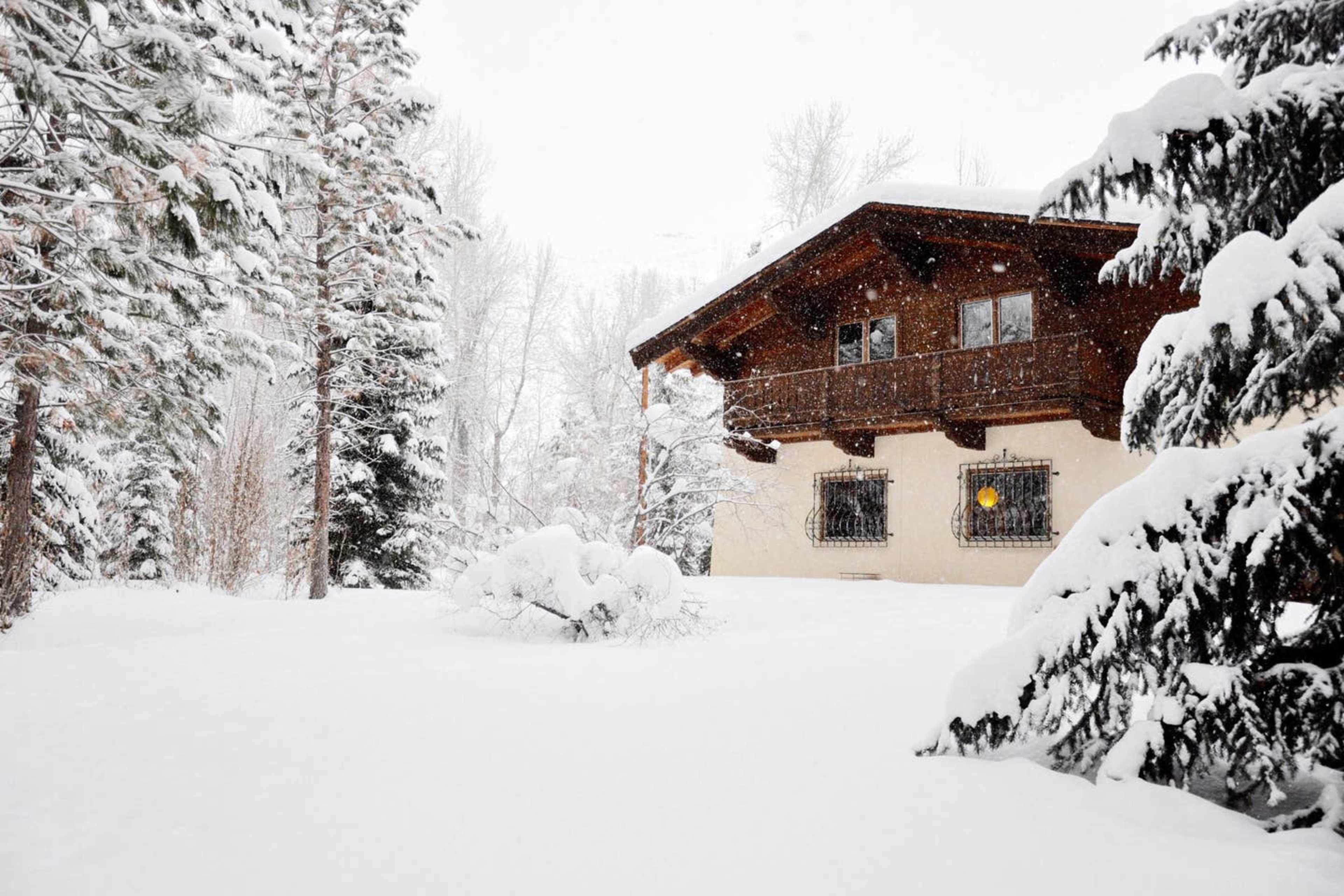 A wooden house with balconies stands in a snowy landscape, surrounded by tall trees and falling snowflakes.