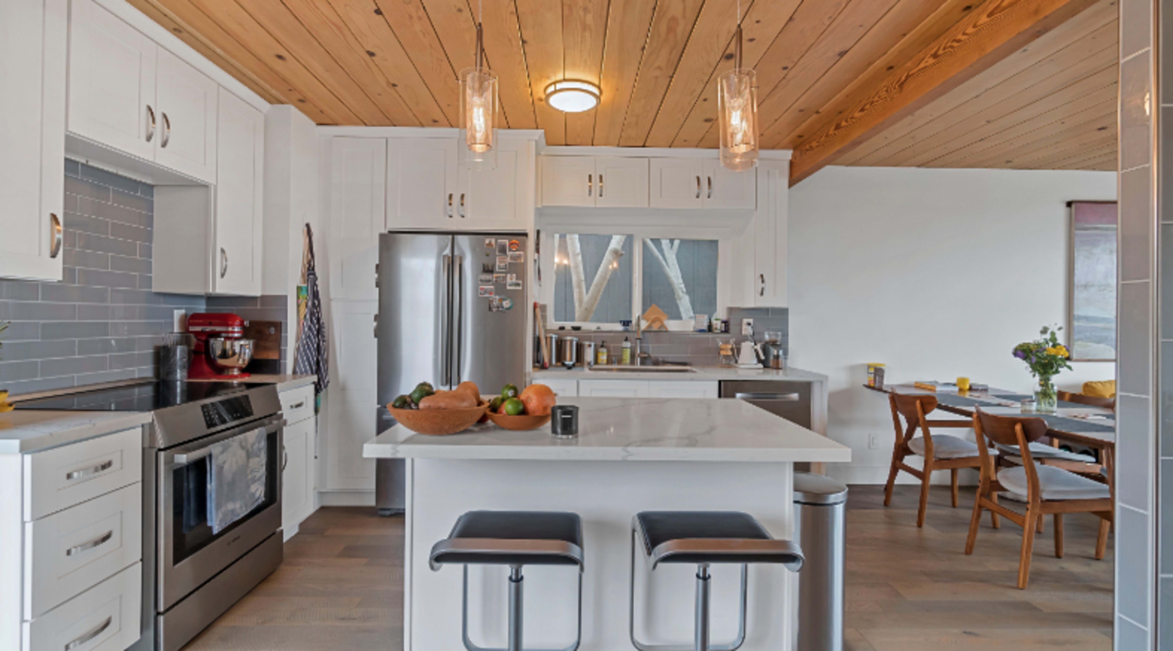 A modern kitchen features white cabinetry, stainless steel appliances, a central island with bar stools, and wooden beams on the ceiling.