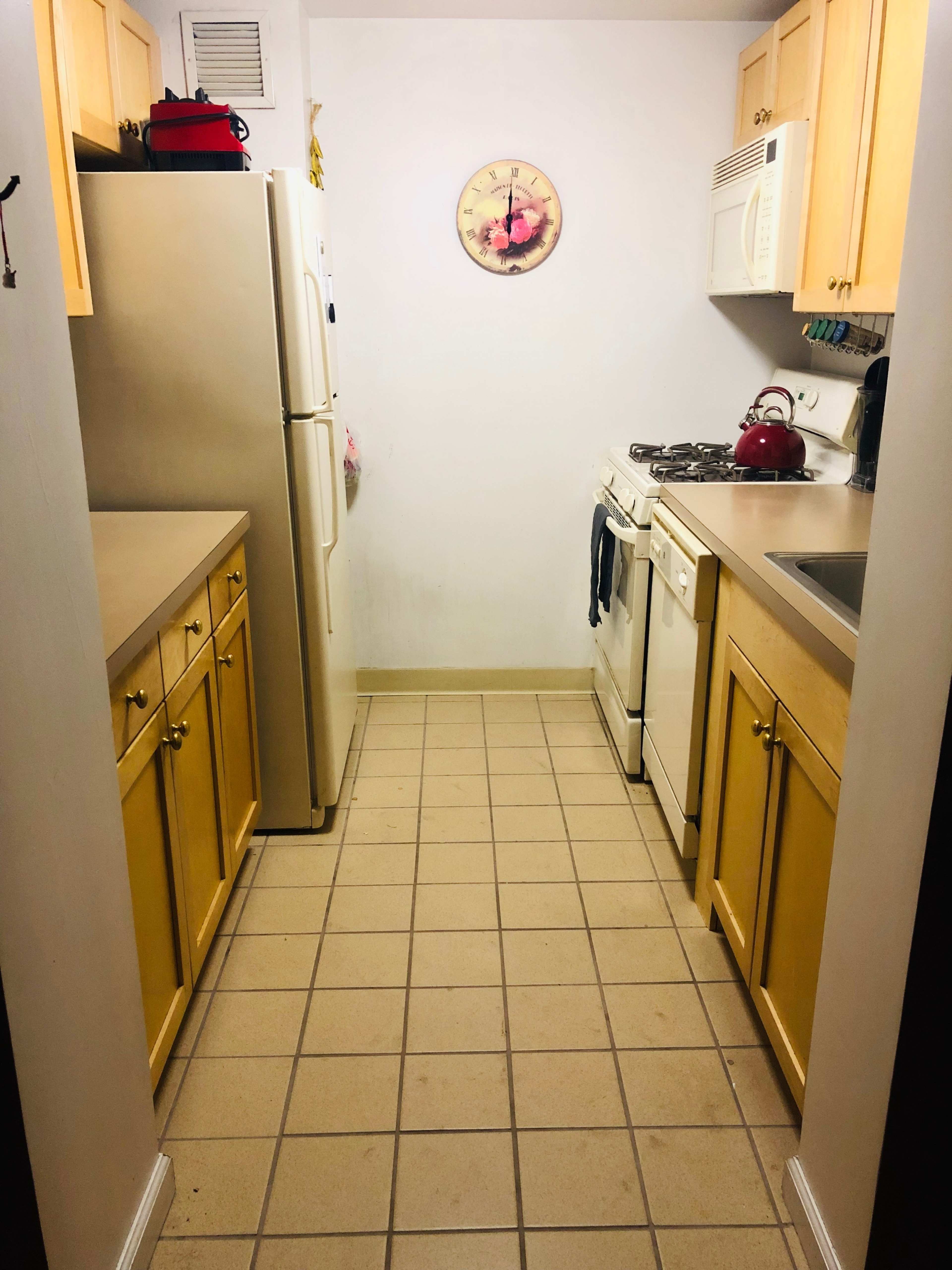 The image shows a narrow kitchen with light wooden cabinets, a stove, sink, and appliances lined along the walls, and a clock on the wall.