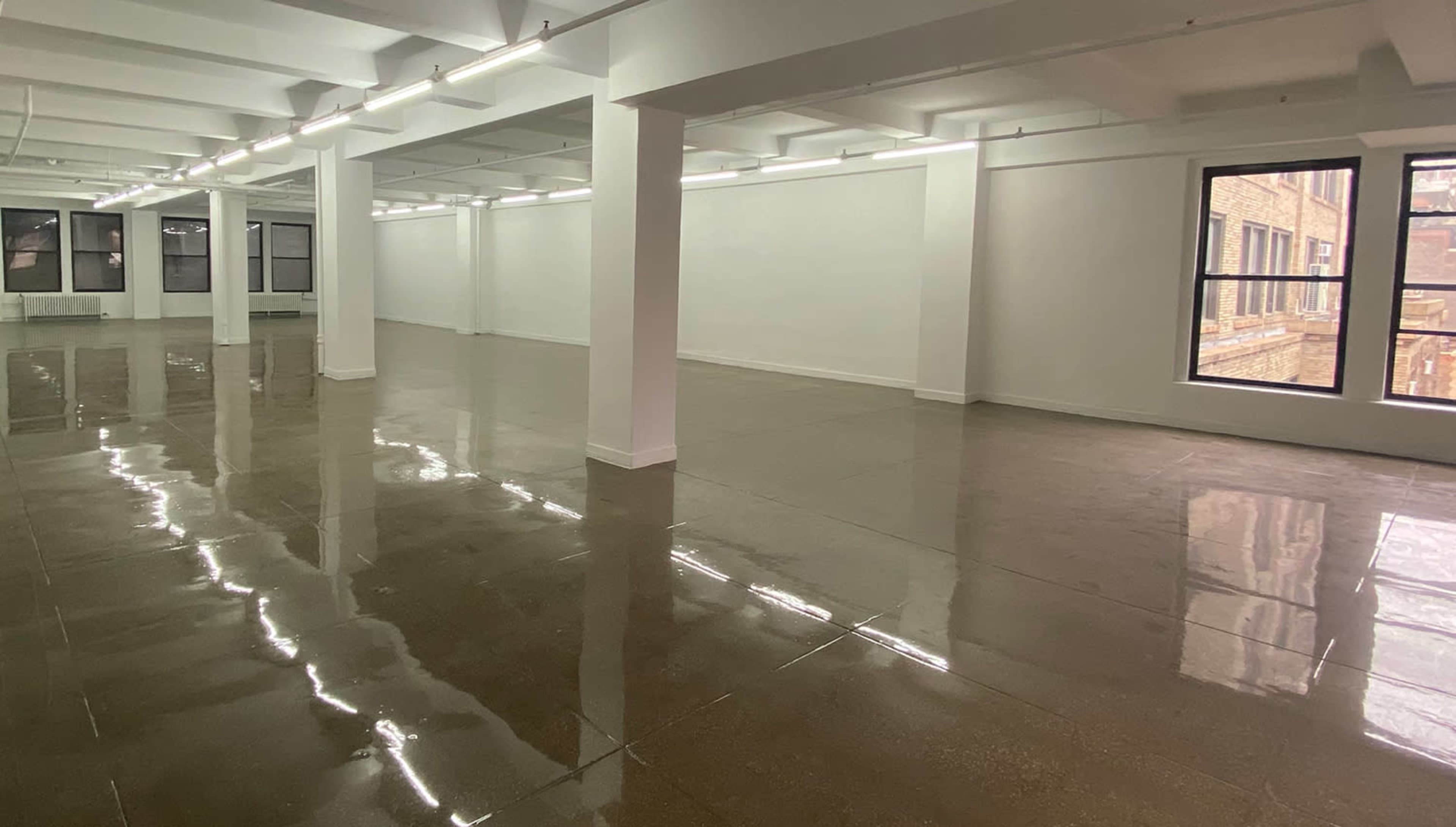 A large, empty room with polished concrete floors and white walls, illuminated by fluorescent lighting.