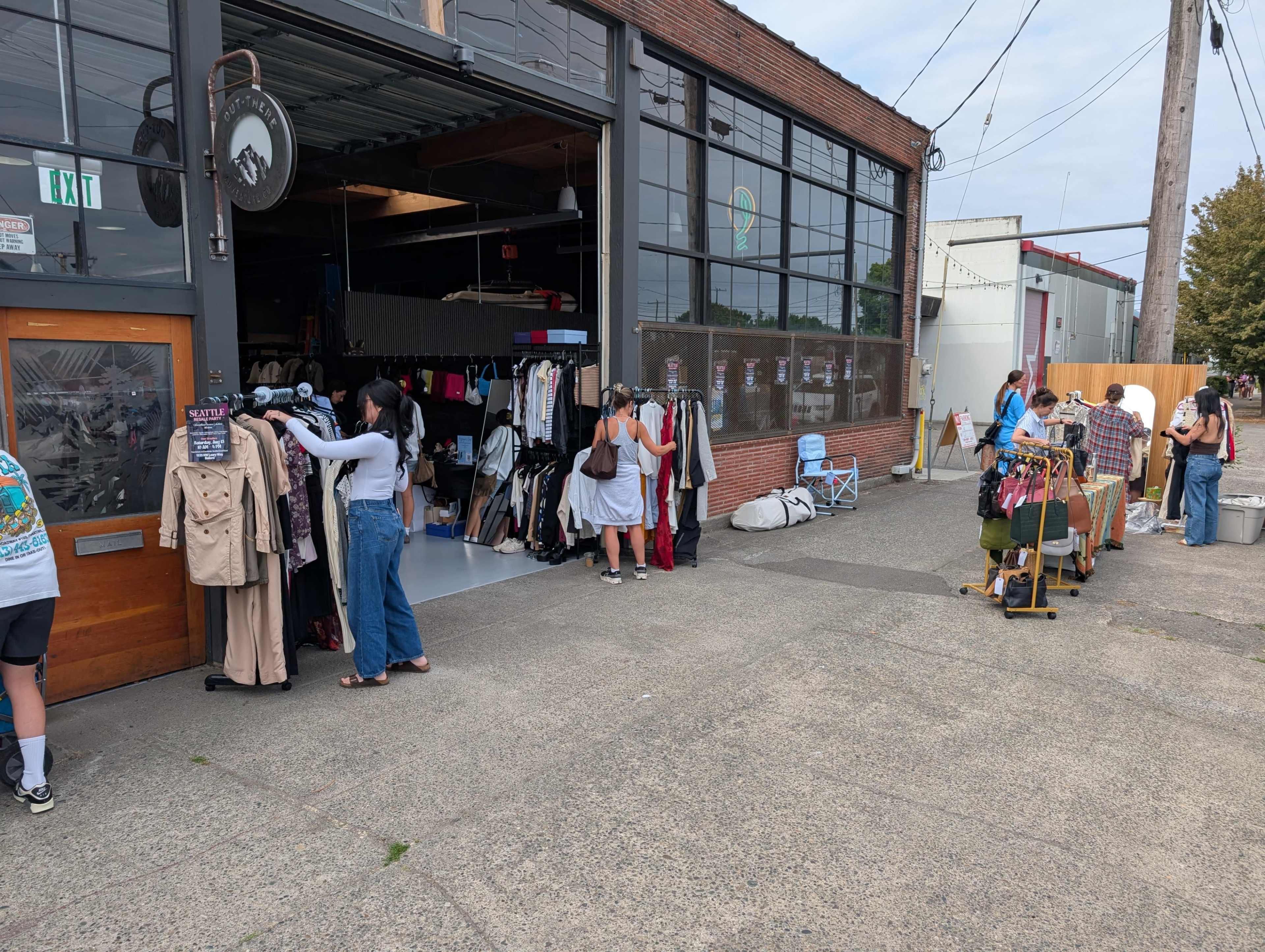 A group of people shops at an outdoor market set up next to a warehouse, with clothing racks and tables displaying various items.
