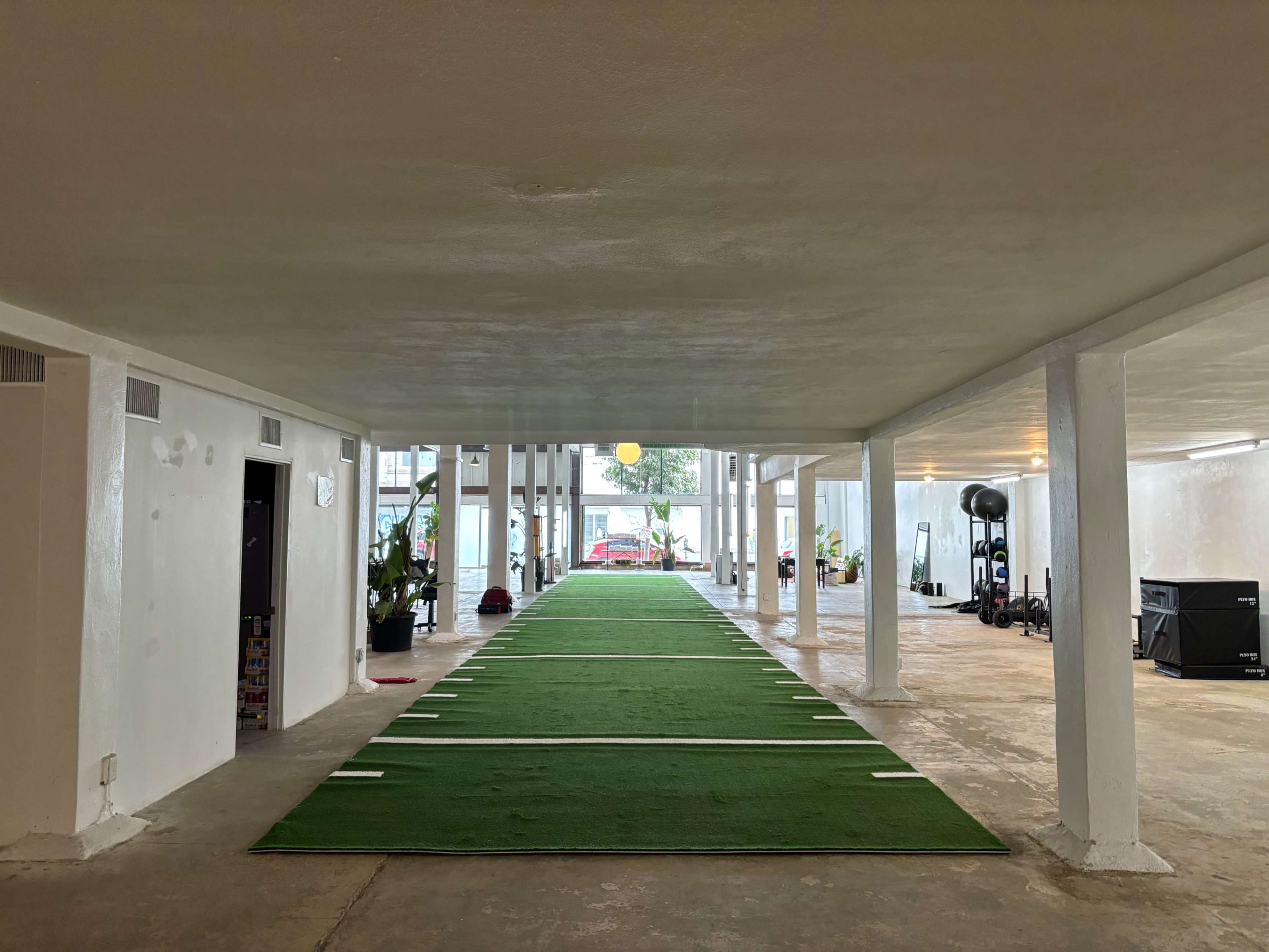 A long indoor corridor with artificial turf lining the center, leading to a distant area with vehicles and fitness equipment on either side.