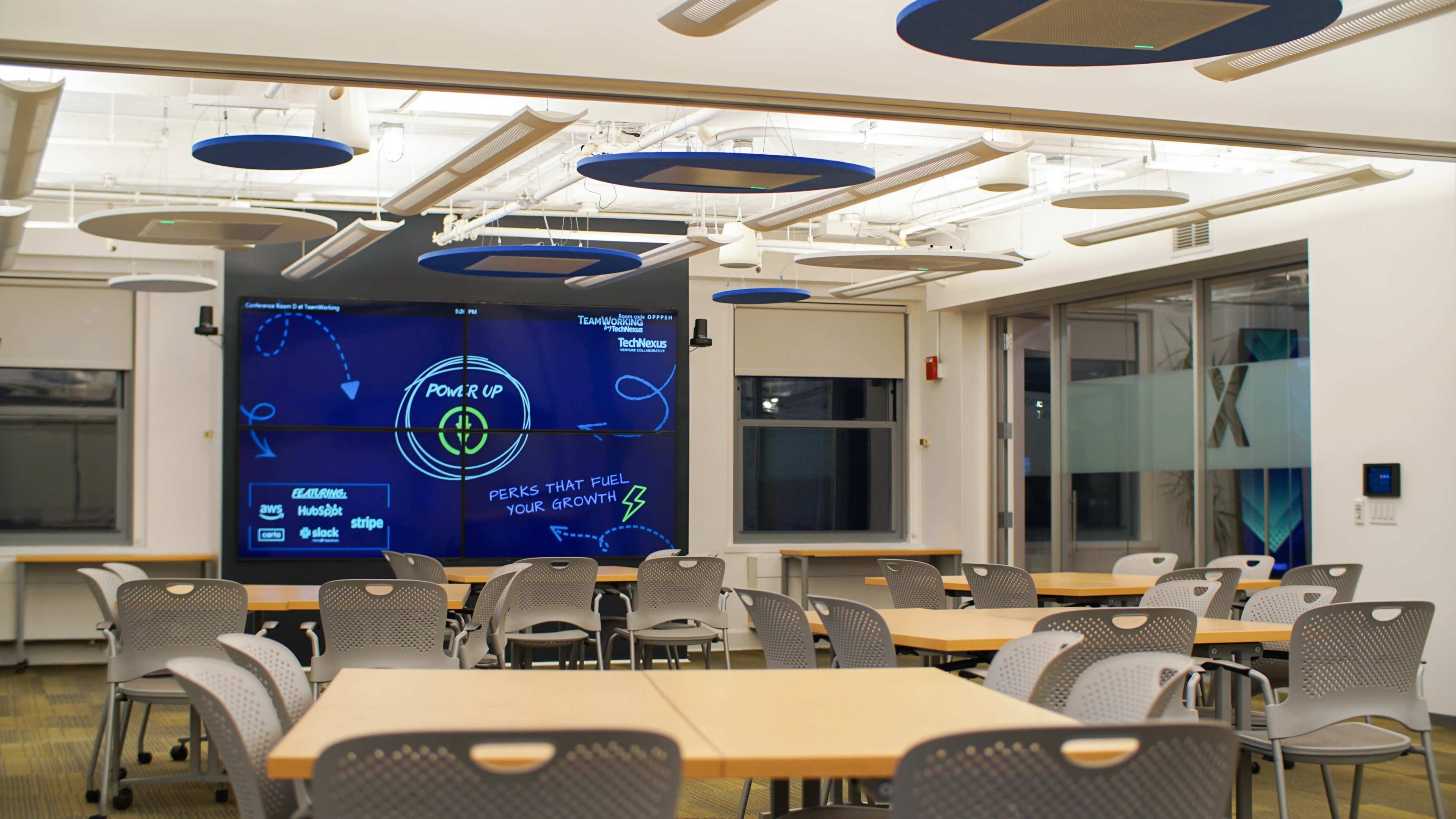 The image shows a modern classroom with rows of tables and chairs, featuring a large screen displaying educational graphics on one wall.
