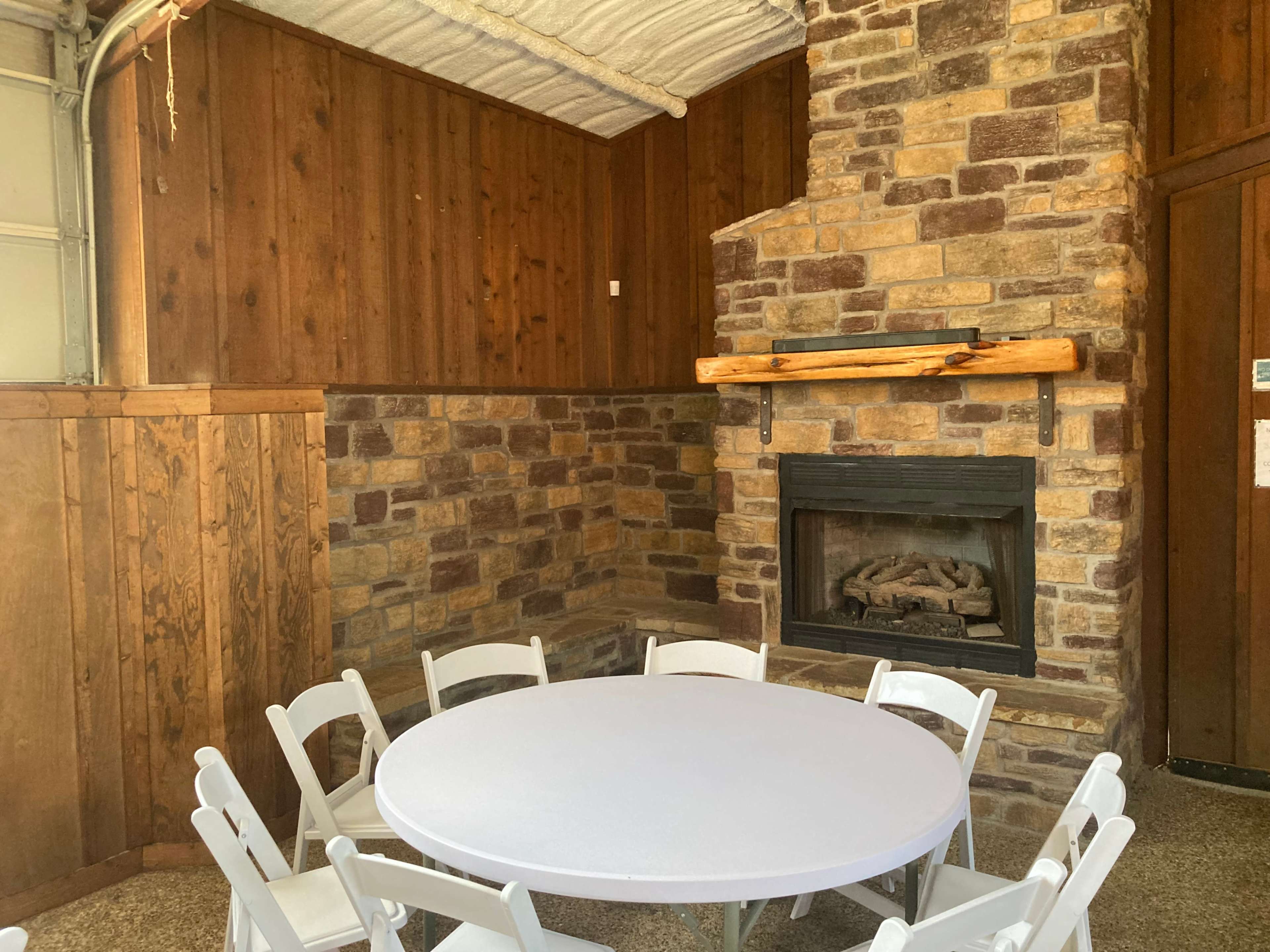 A cozy seating area with a round white table surrounded by white chairs, complemented by a stone fireplace and wooden paneling on the walls.