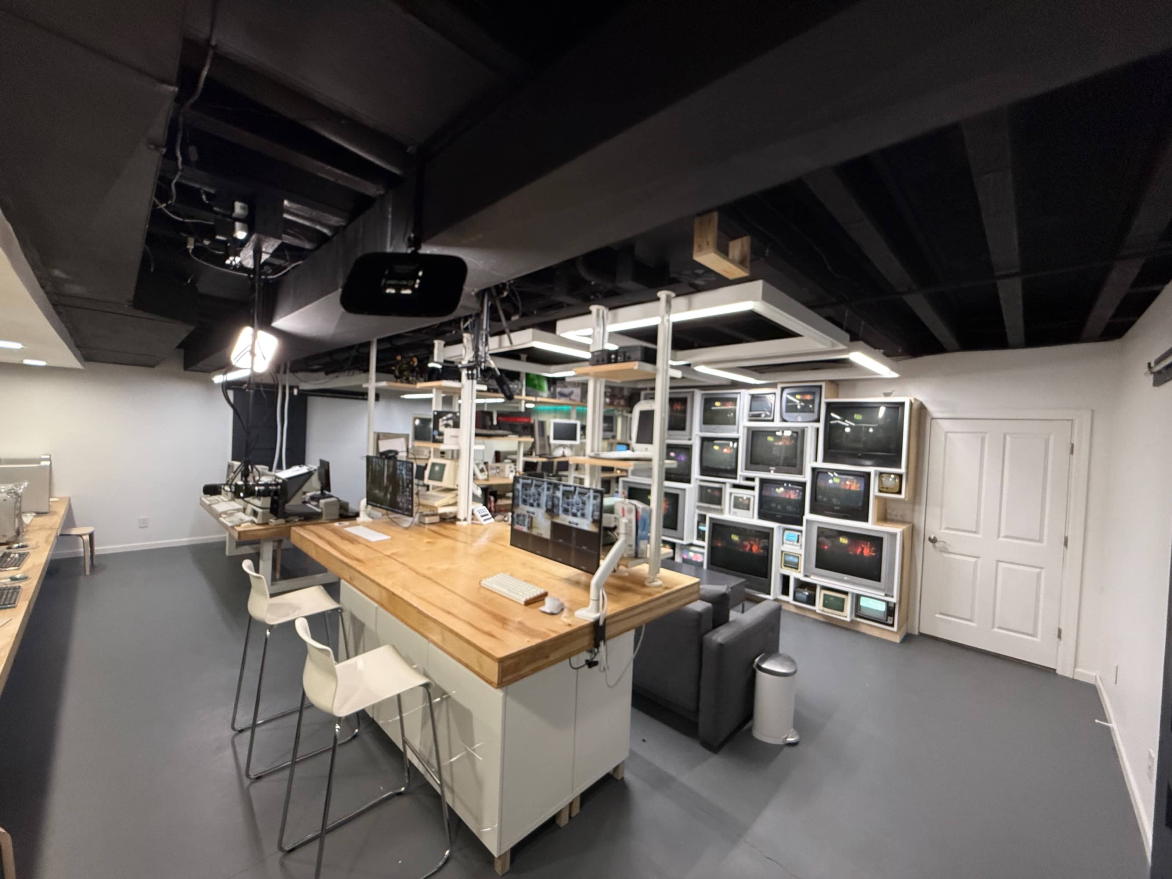 The image shows a modern office space featuring a wooden countertop, multiple computer screens, and a seating area with a black sofa against a wall of displays.