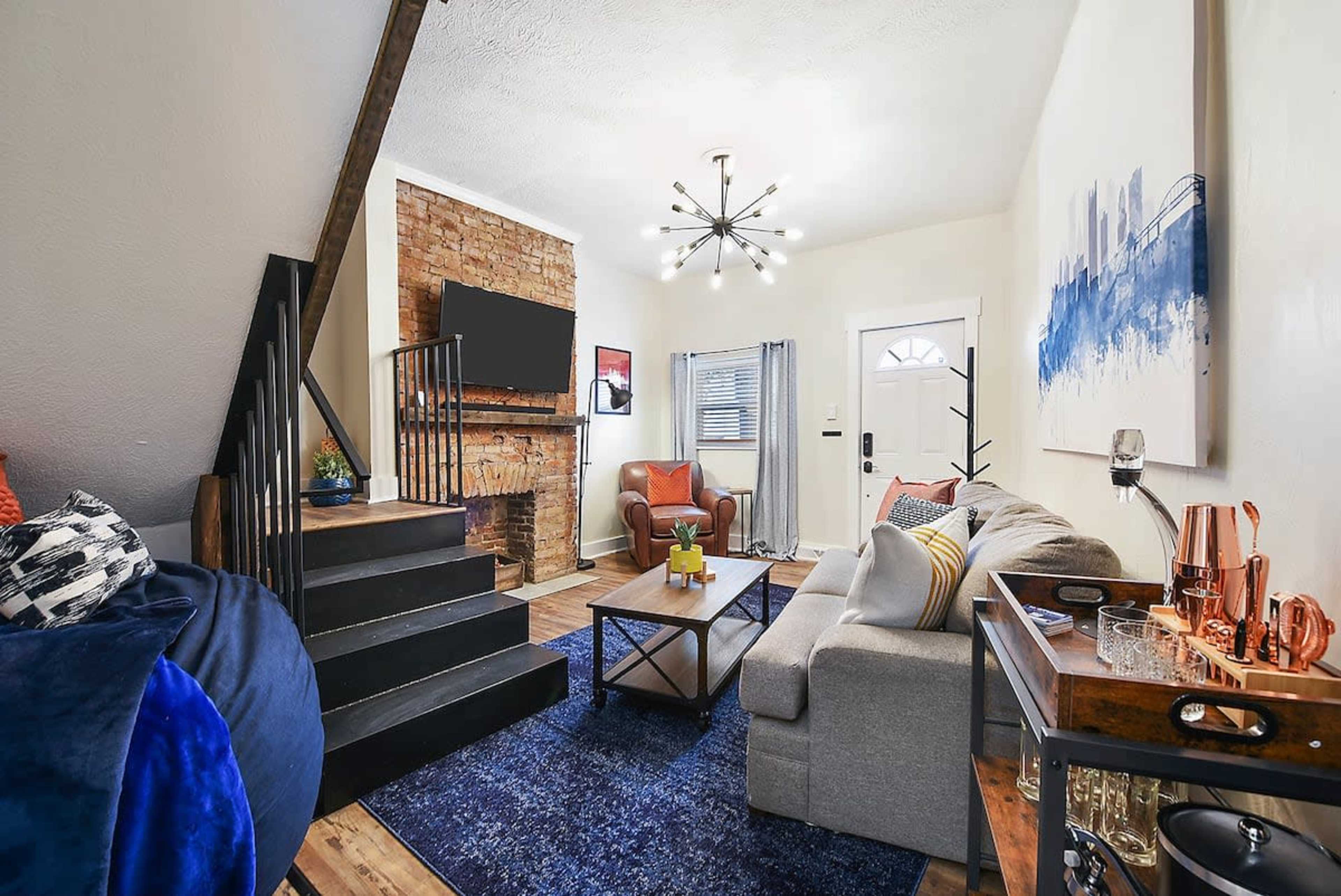 A cozy living room features a staircase, a brick accent wall with a television, a sofa, an armchair, and a coffee table on a blue area rug.