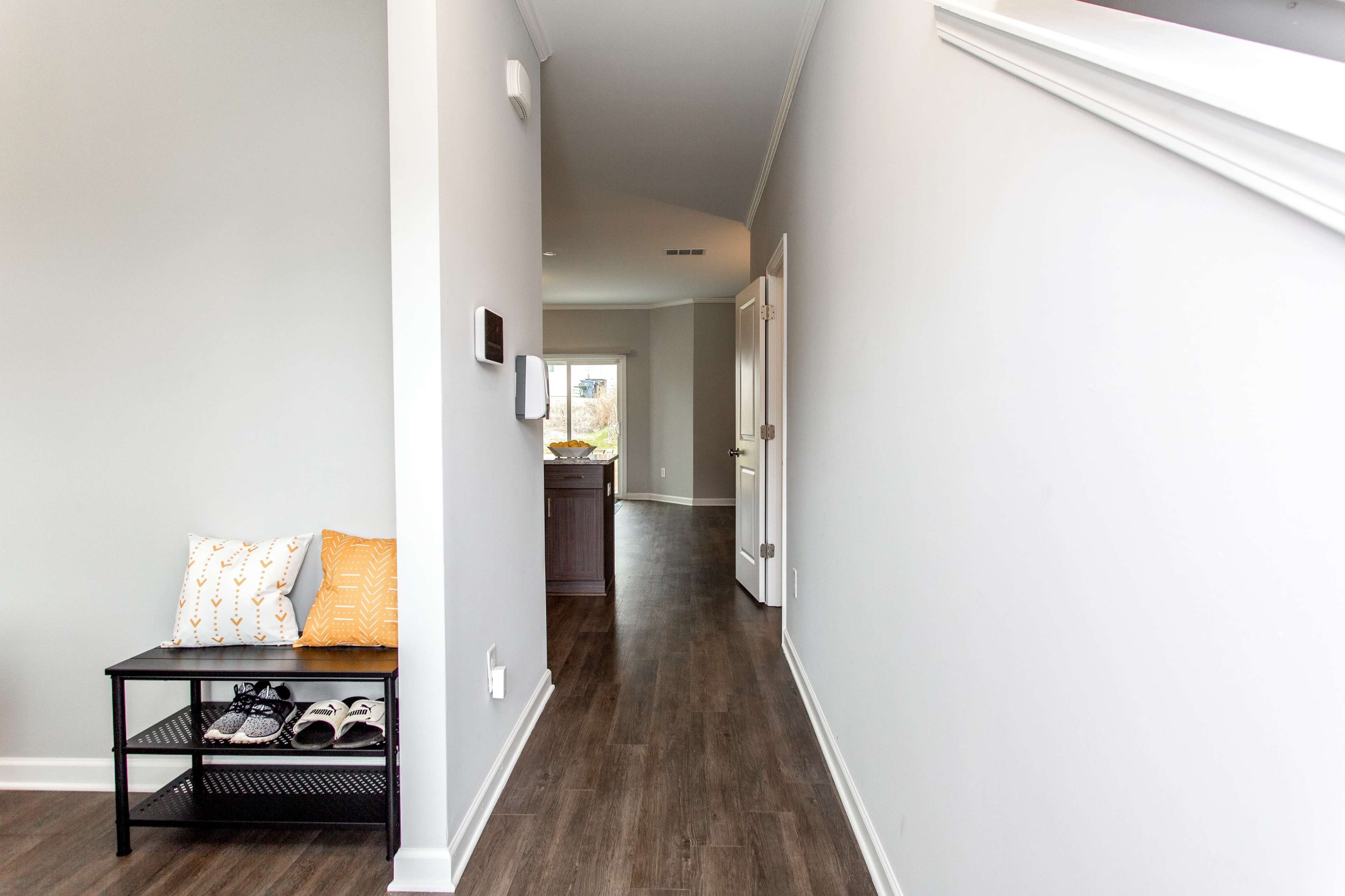 A narrow hallway with a bench holding decorative cushions and a pair of shoes, leading towards an open kitchen area in the background.