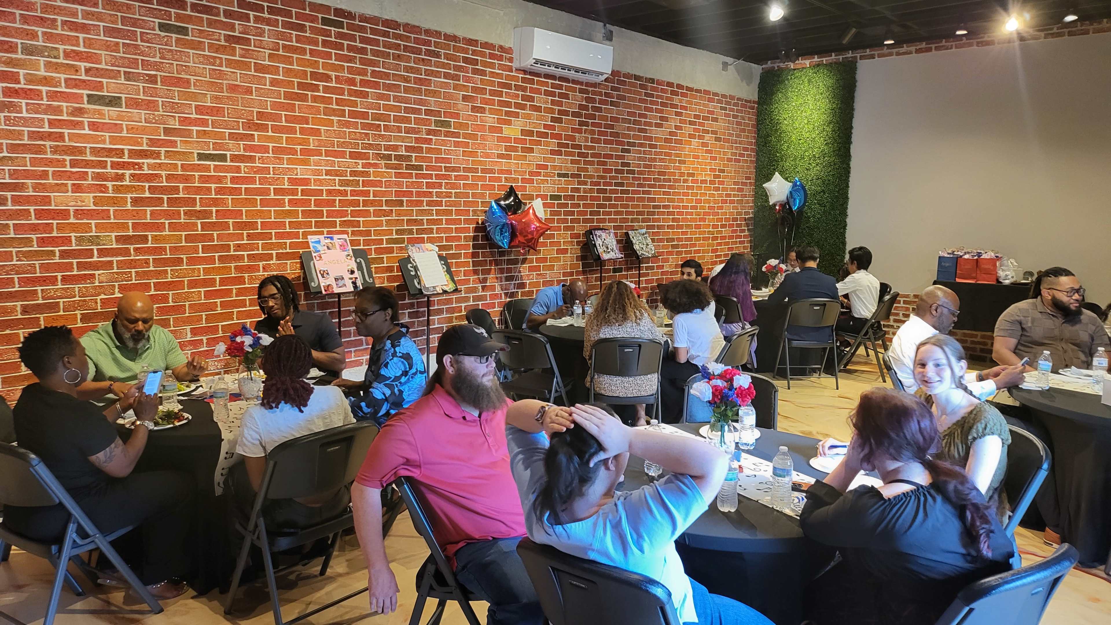 The image shows a gathering in a spacious room with people seated at round tables, engaged in conversation, and decorative elements including balloons and framed photos displayed on the walls.