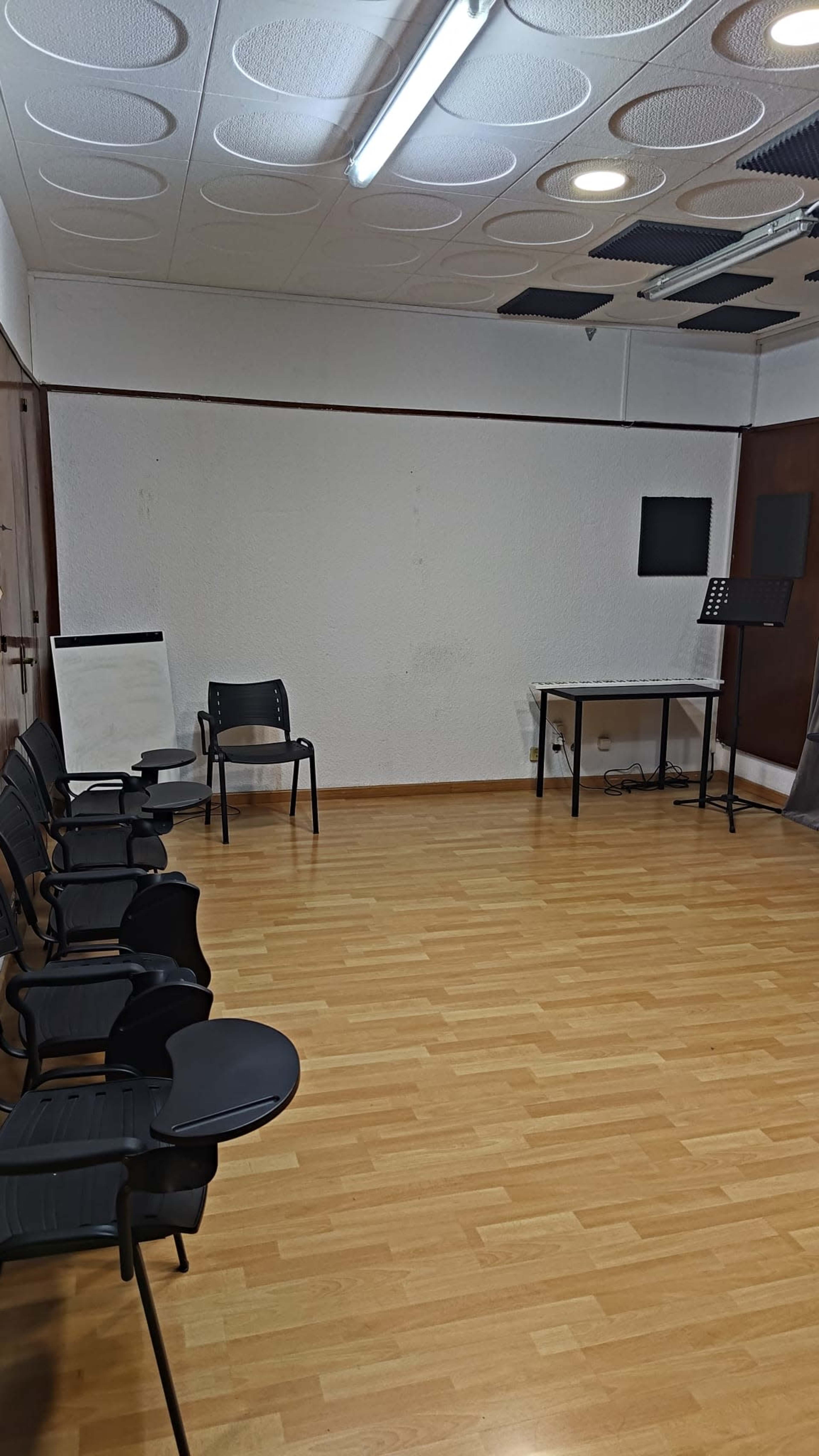 The image shows a spacious, empty room with wooden flooring, several black chairs, a flip chart, a table, and two mounted screens on the walls.