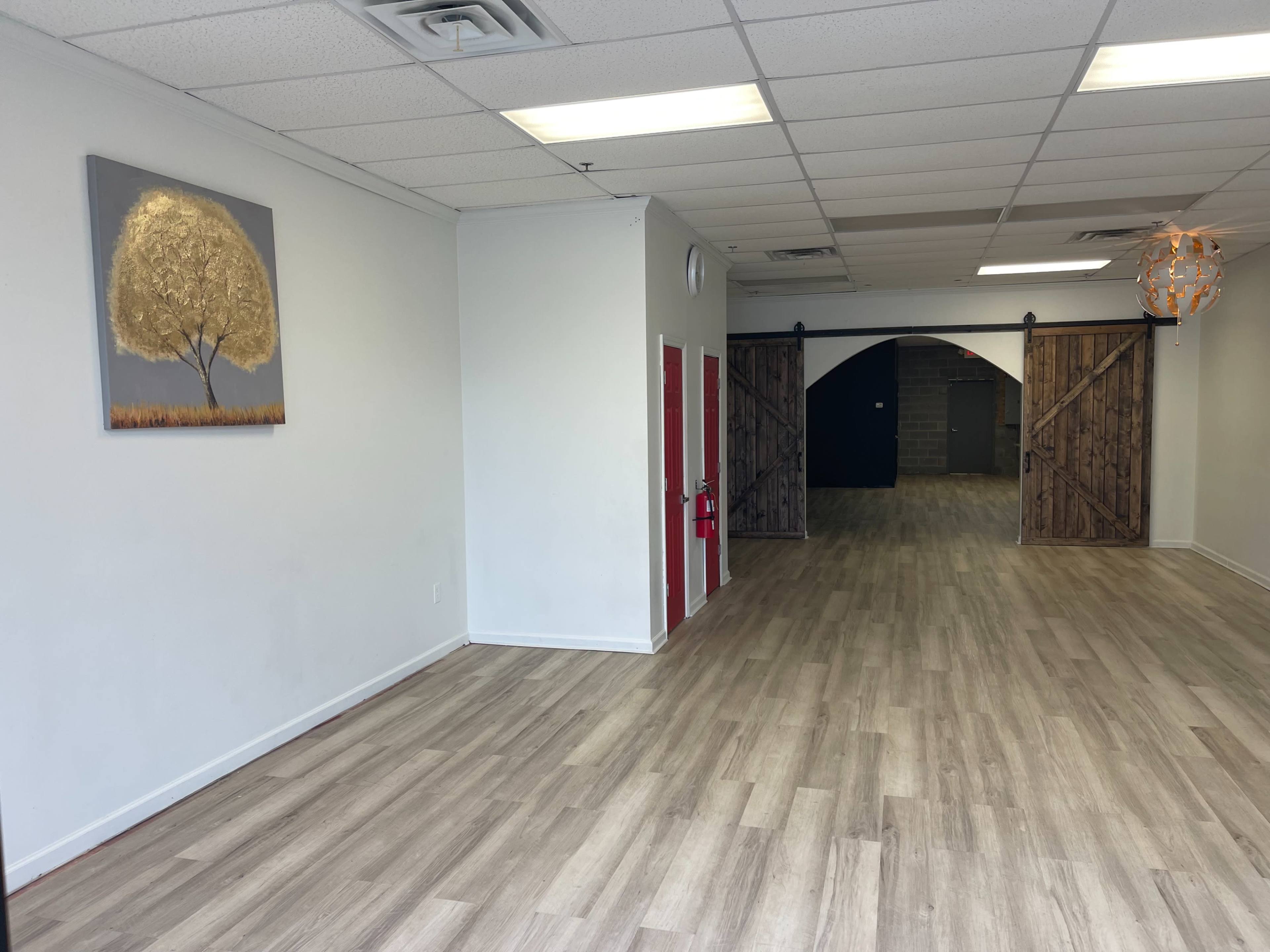 The image shows an empty interior space with wood flooring, a painting of a tree on the left wall, and double doors made of wood leading to a darker area at the back.