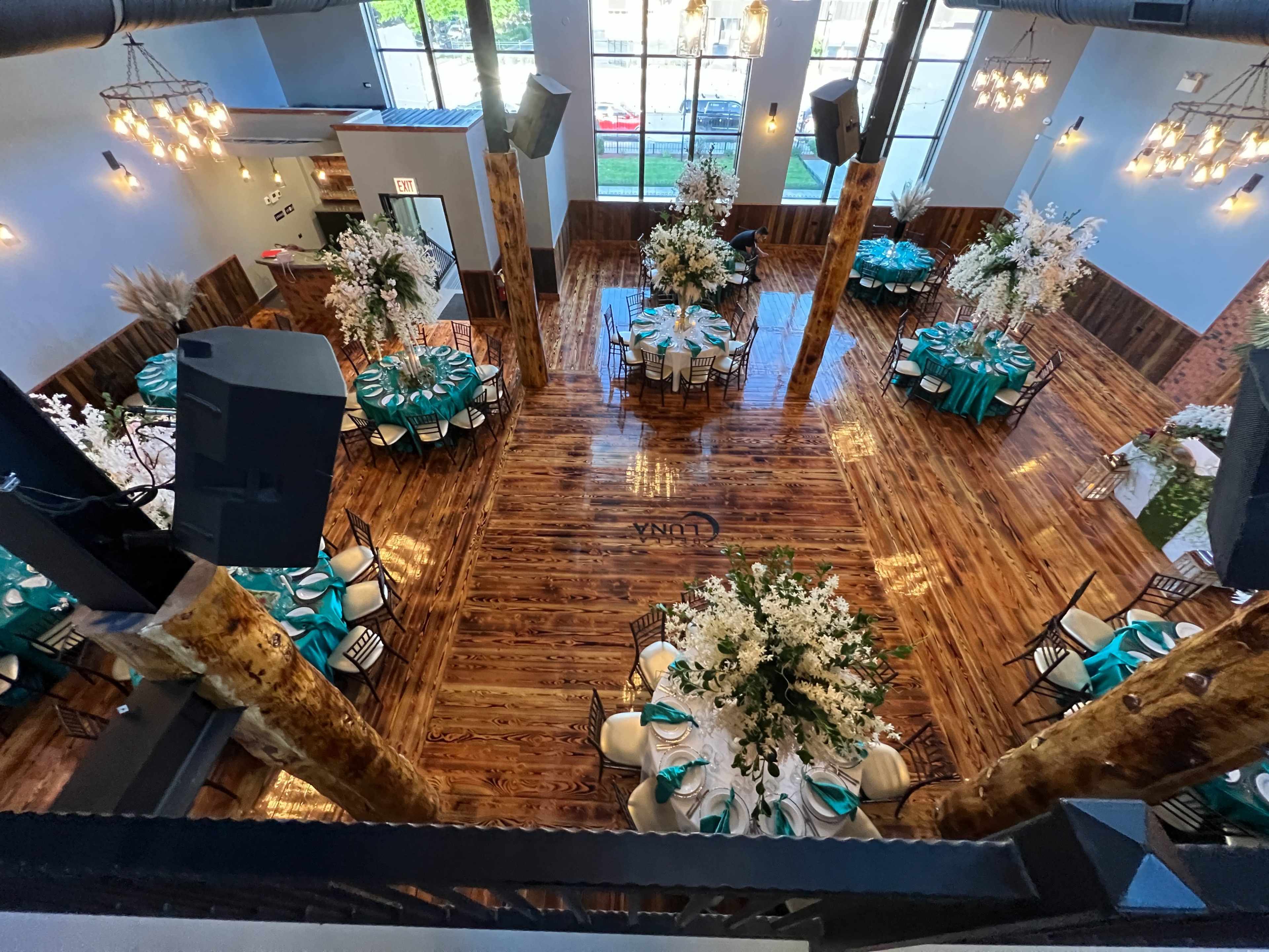 An elegantly arranged event space with tables set and decorated with turquoise and white accents, surrounded by tall floral centerpieces and wooden pillars.
