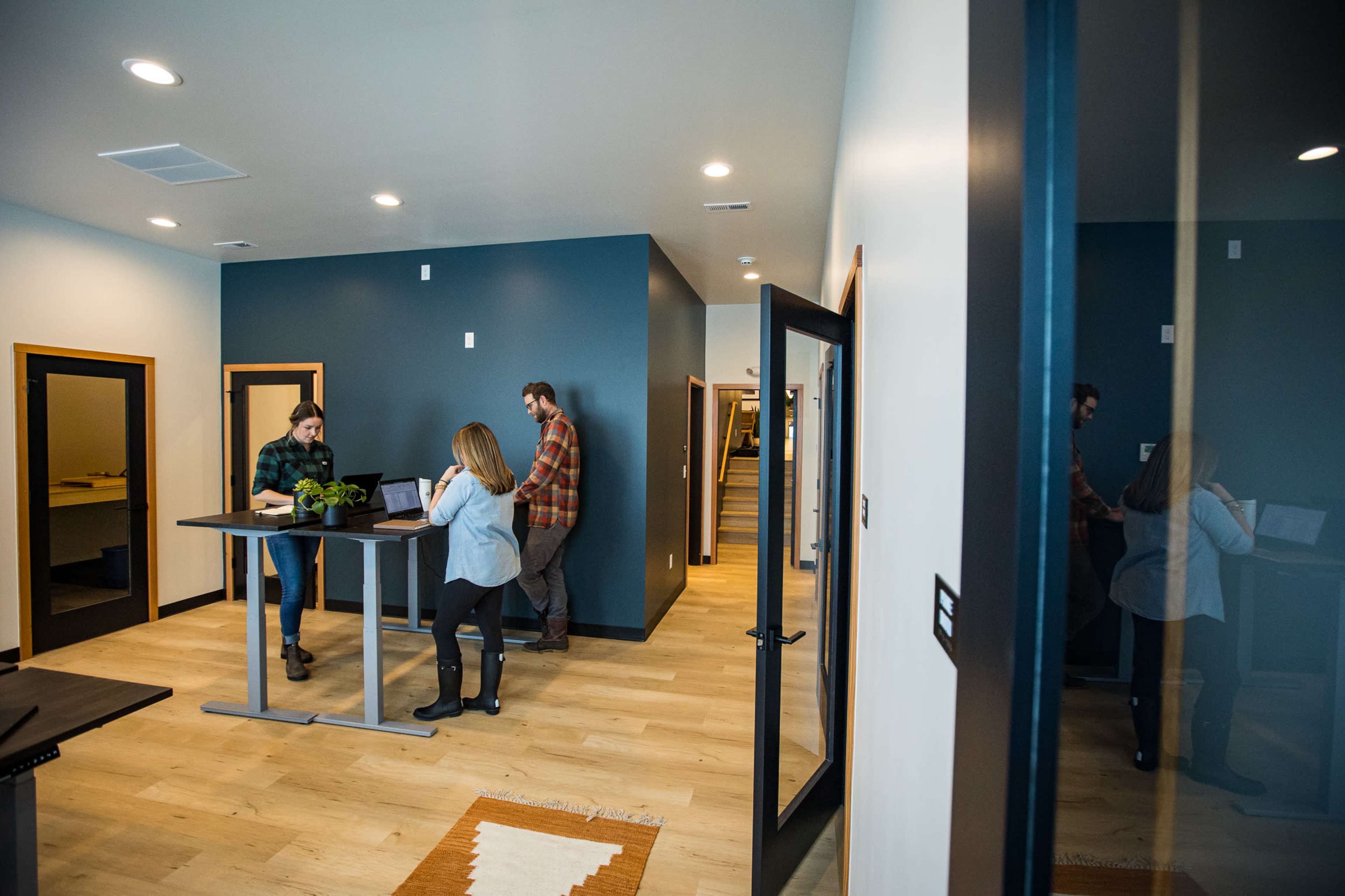 The image shows a modern office space with three people engaging in work at standing desks, surrounded by neutral-colored walls and wooden flooring.