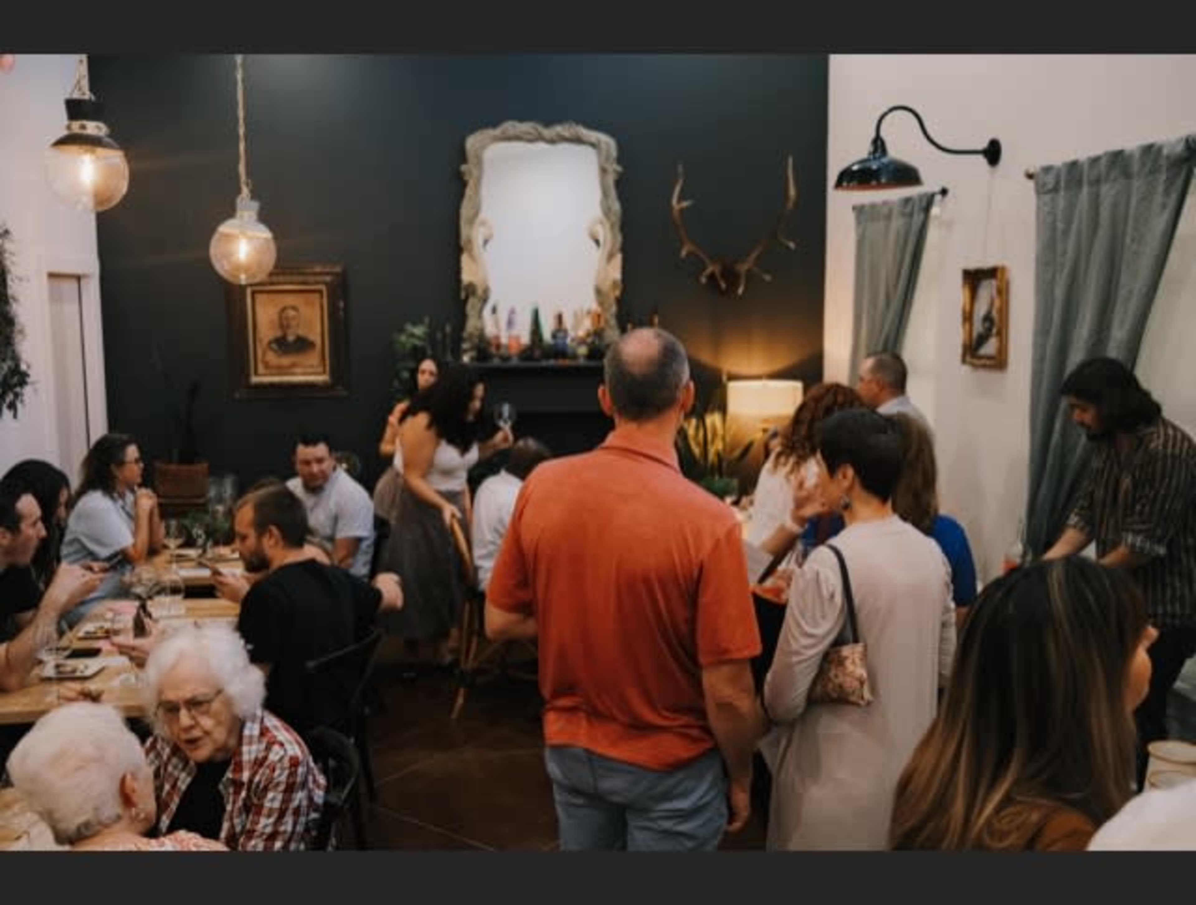 A diverse group of people mingles at a dinner party in a warmly decorated room with a large mirror and various decorative items on the mantel.