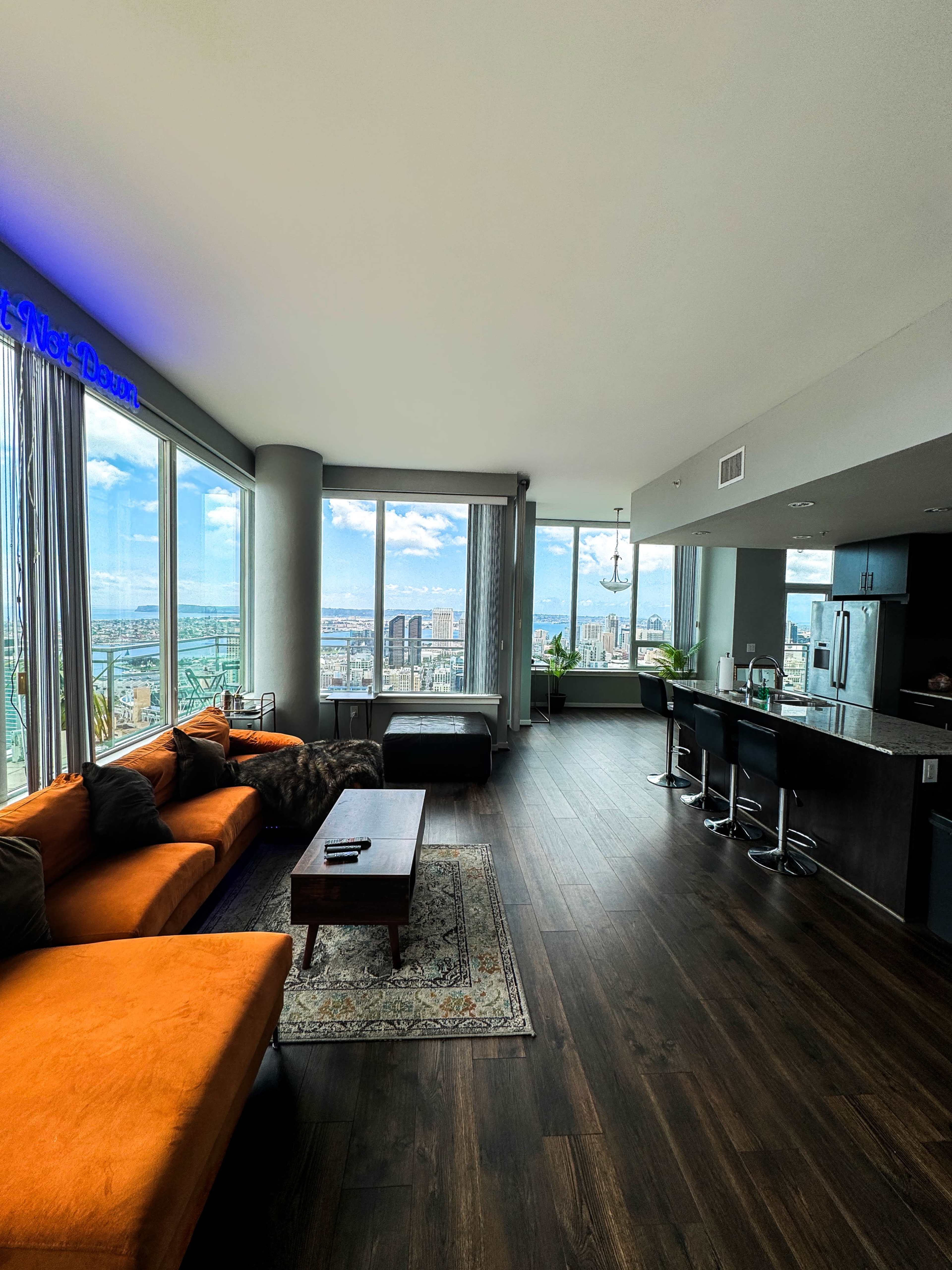 A modern high-rise apartment features an open living area with large windows showcasing a city view, orange couch, and a kitchen bar with stools.