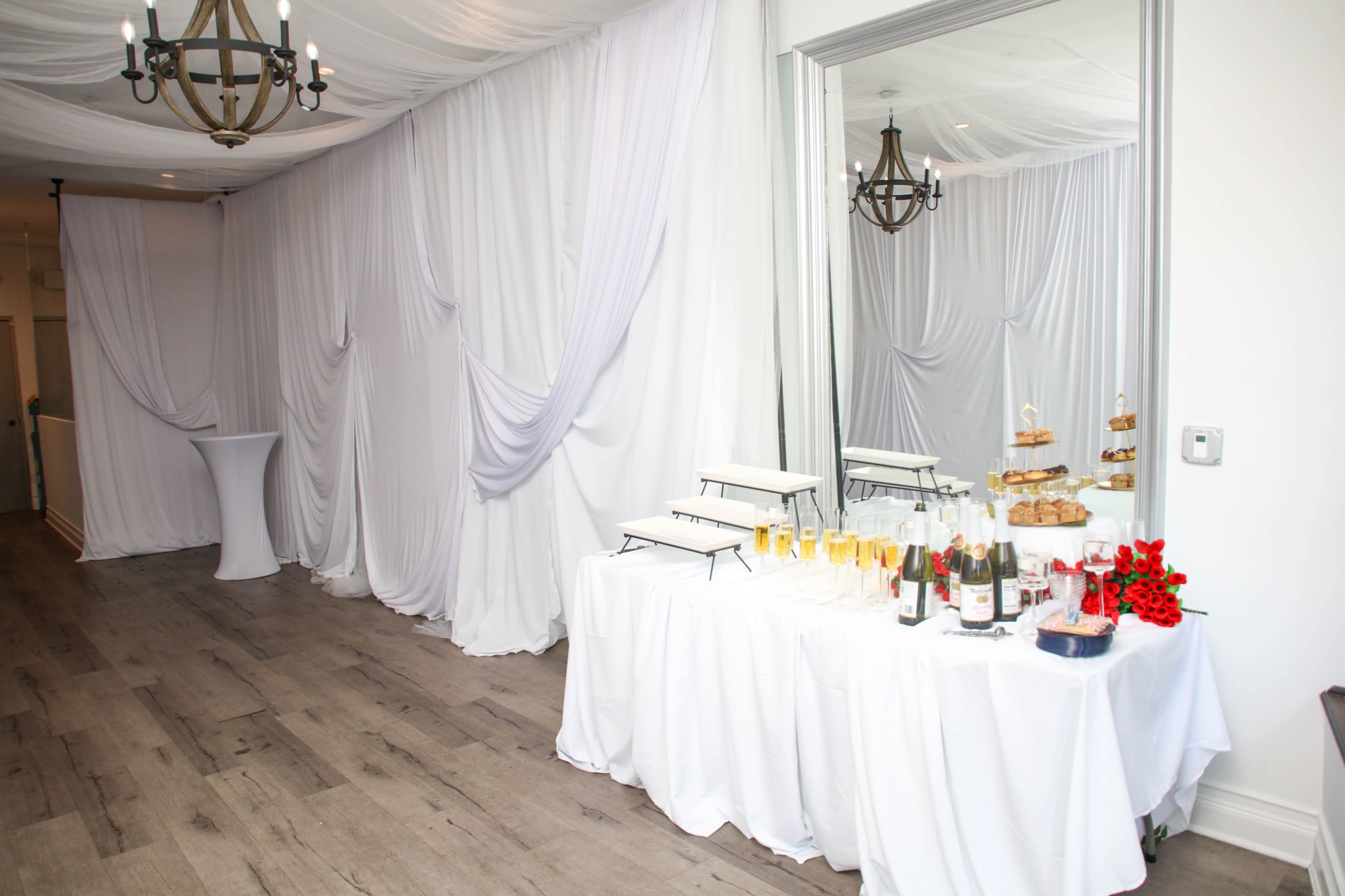 The image shows a decorated event space featuring draped white fabric, a table with drinks and snacks, and a large mirror reflecting the setting.