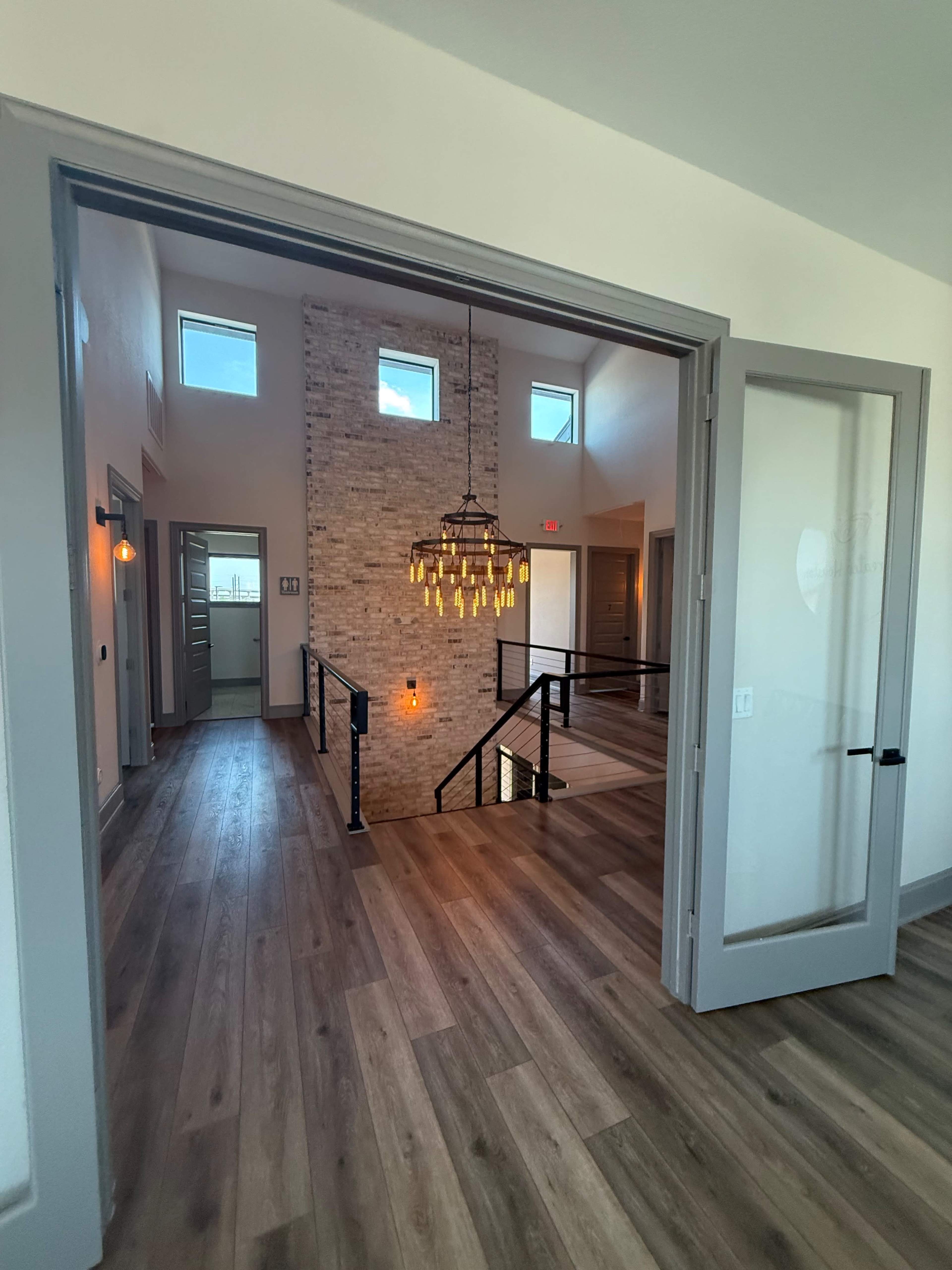 The image shows a modern interior space with a wooden floor, exposed brick wall, and a hanging light fixture, leading to rooms with large windows.
