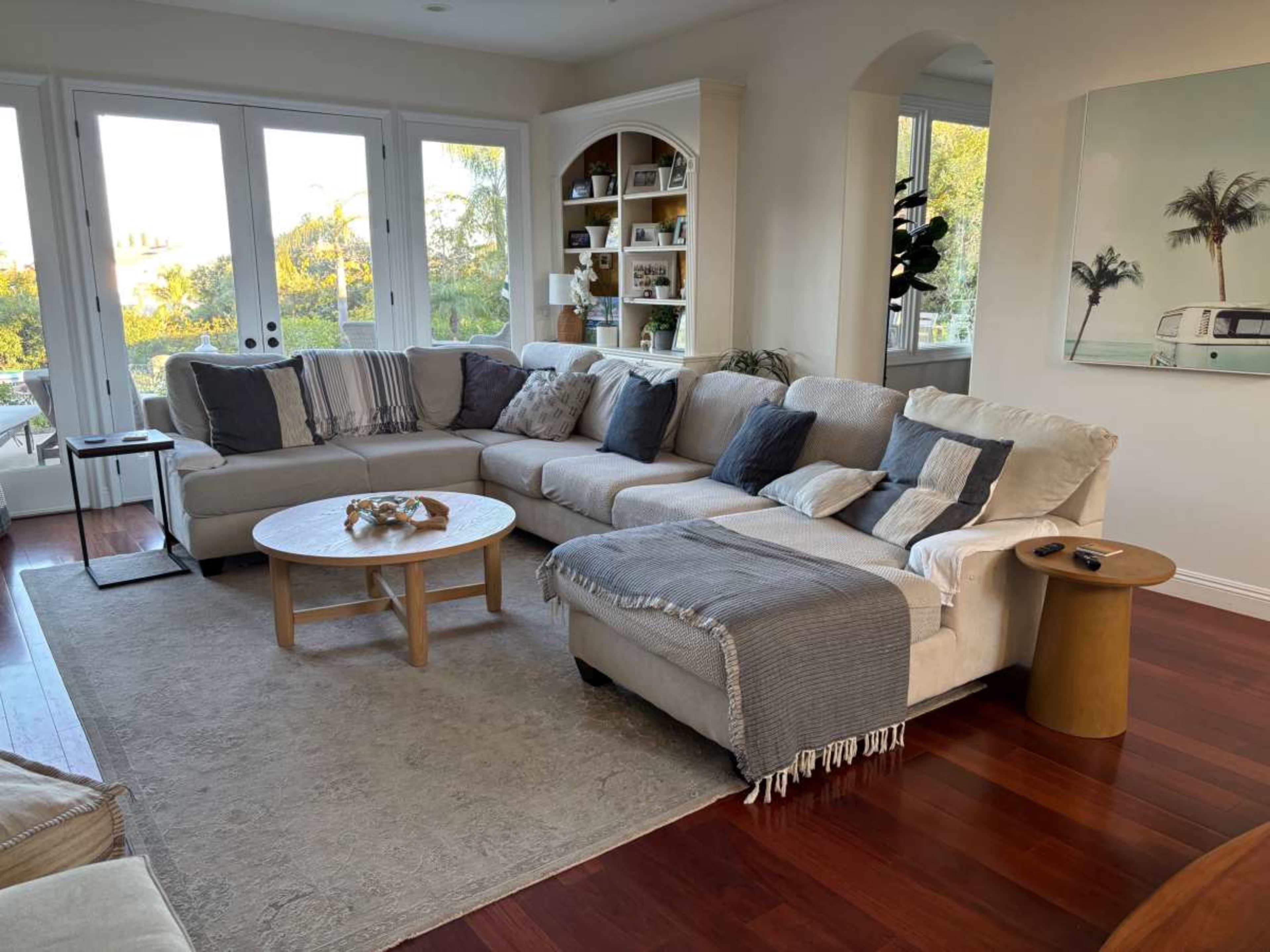 The image shows a cozy living room with a large sectional sofa, a round coffee table, and large windows that let in natural light.