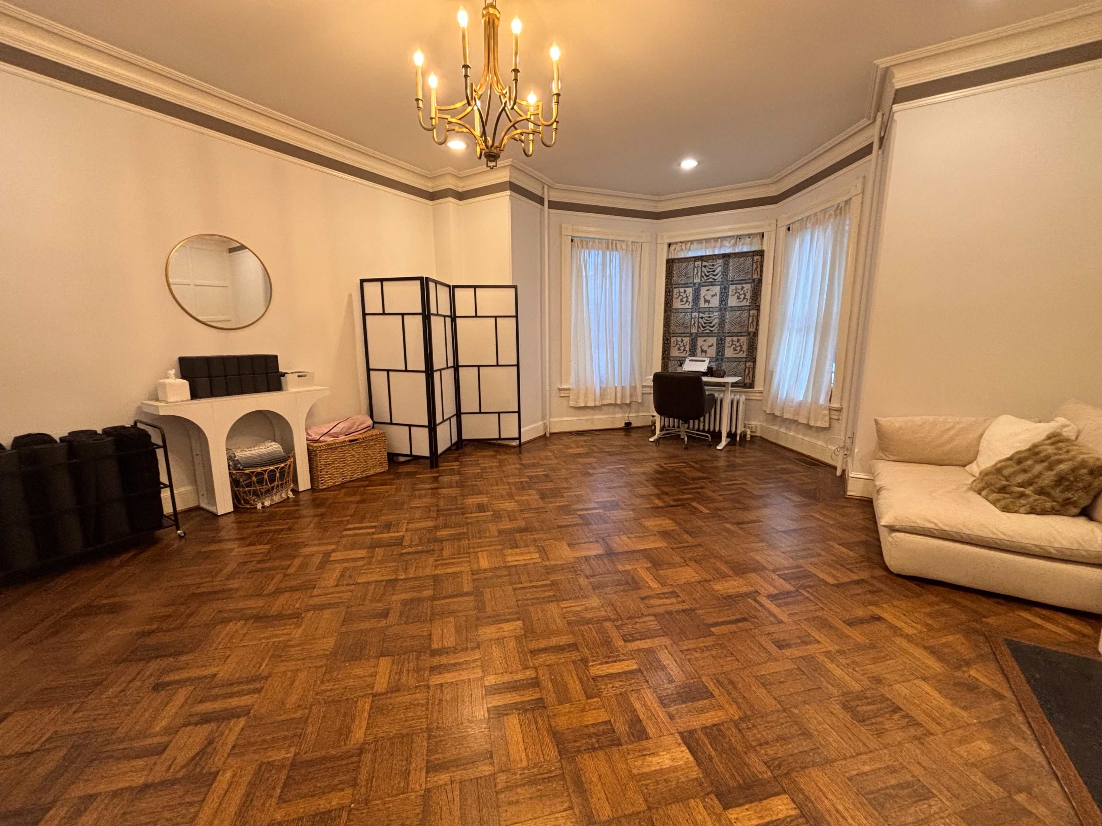 The image shows a spacious room with wooden parquet flooring, a chandelier, large windows with curtains, and a combination of furniture including a sofa, a folding screen, and a desk.