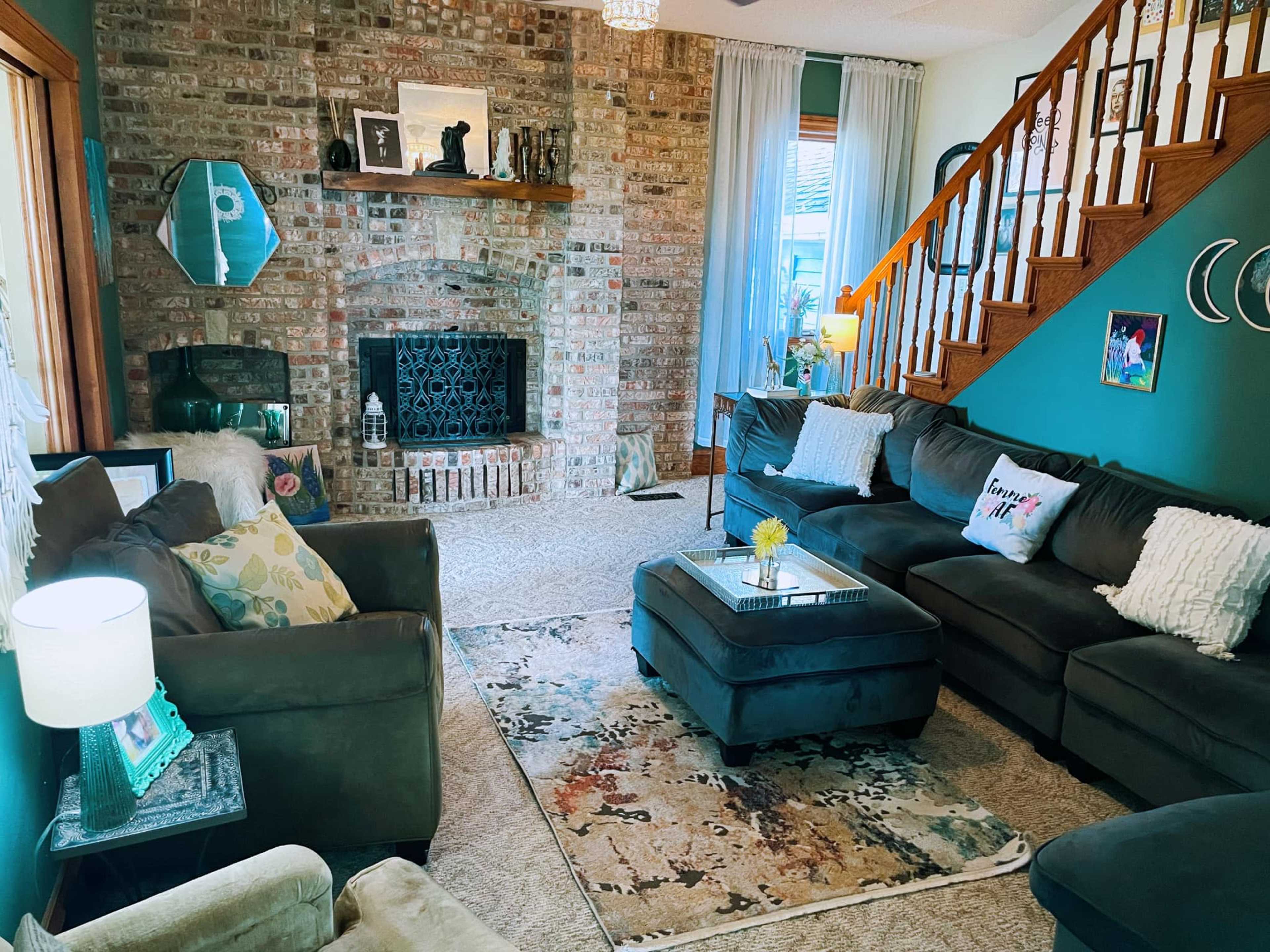 A cozy living room featuring a brick fireplace, a mix of dark-colored sofas, a decorative rug, and a staircase in the background.