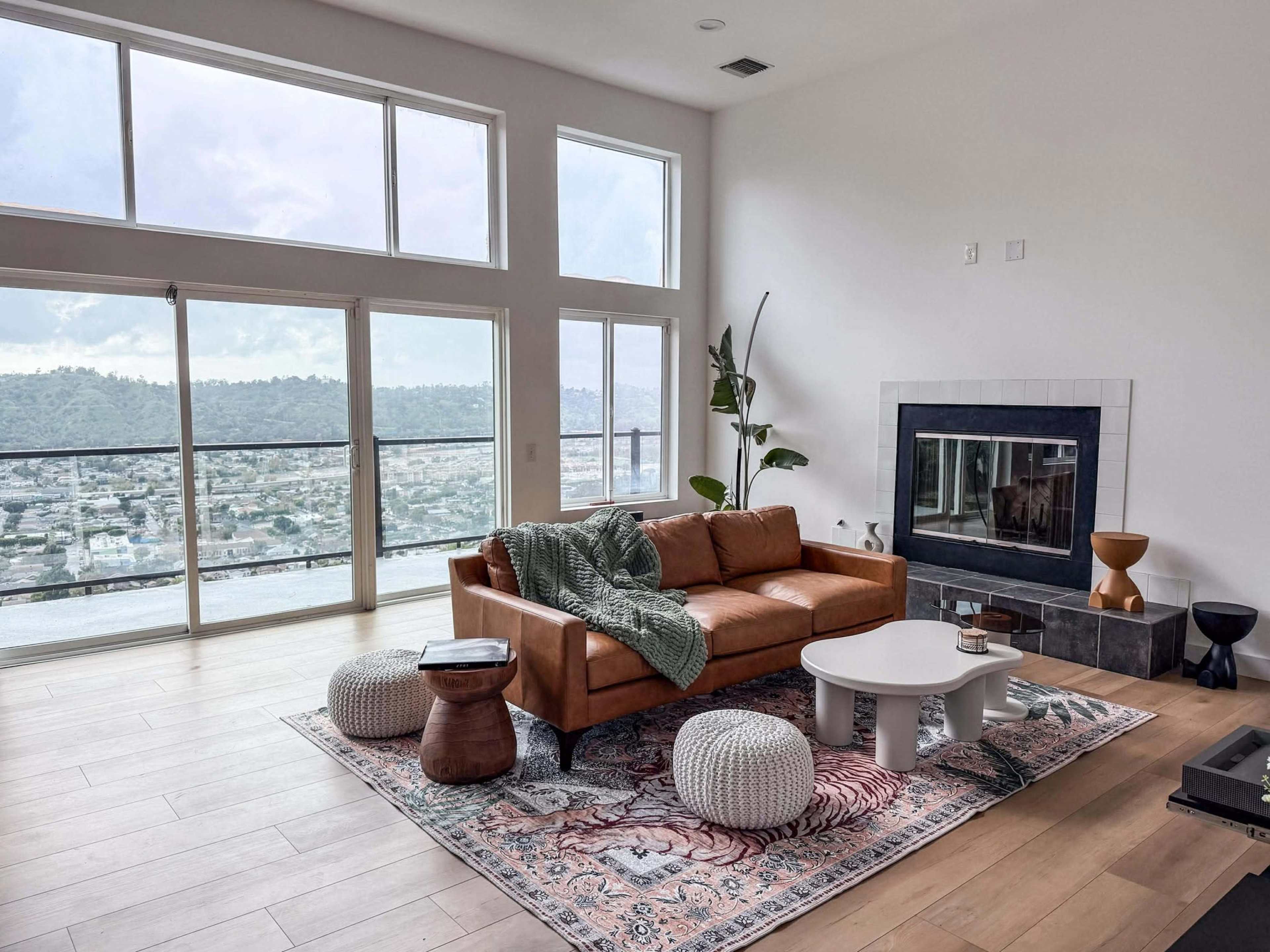 The image shows a spacious living room with large windows offering a view of a hillside cityscape, featuring a brown leather sofa, a white coffee table, decorative poufs, a patterned area rug, and a fireplace.