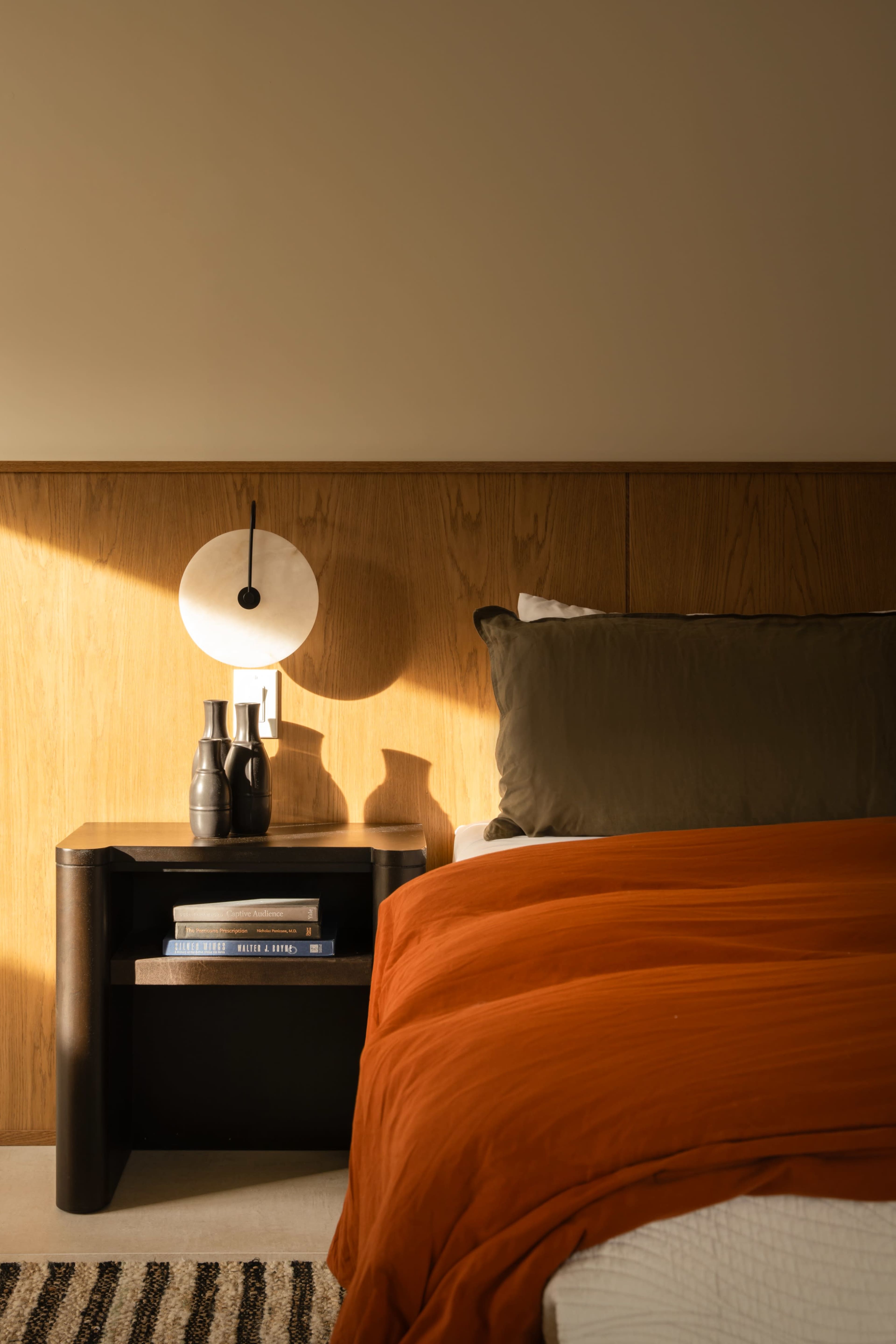 A bedside table with books and decorative vases stands next to a bed with an orange comforter and a dark green pillow, illuminated by soft sunlight.