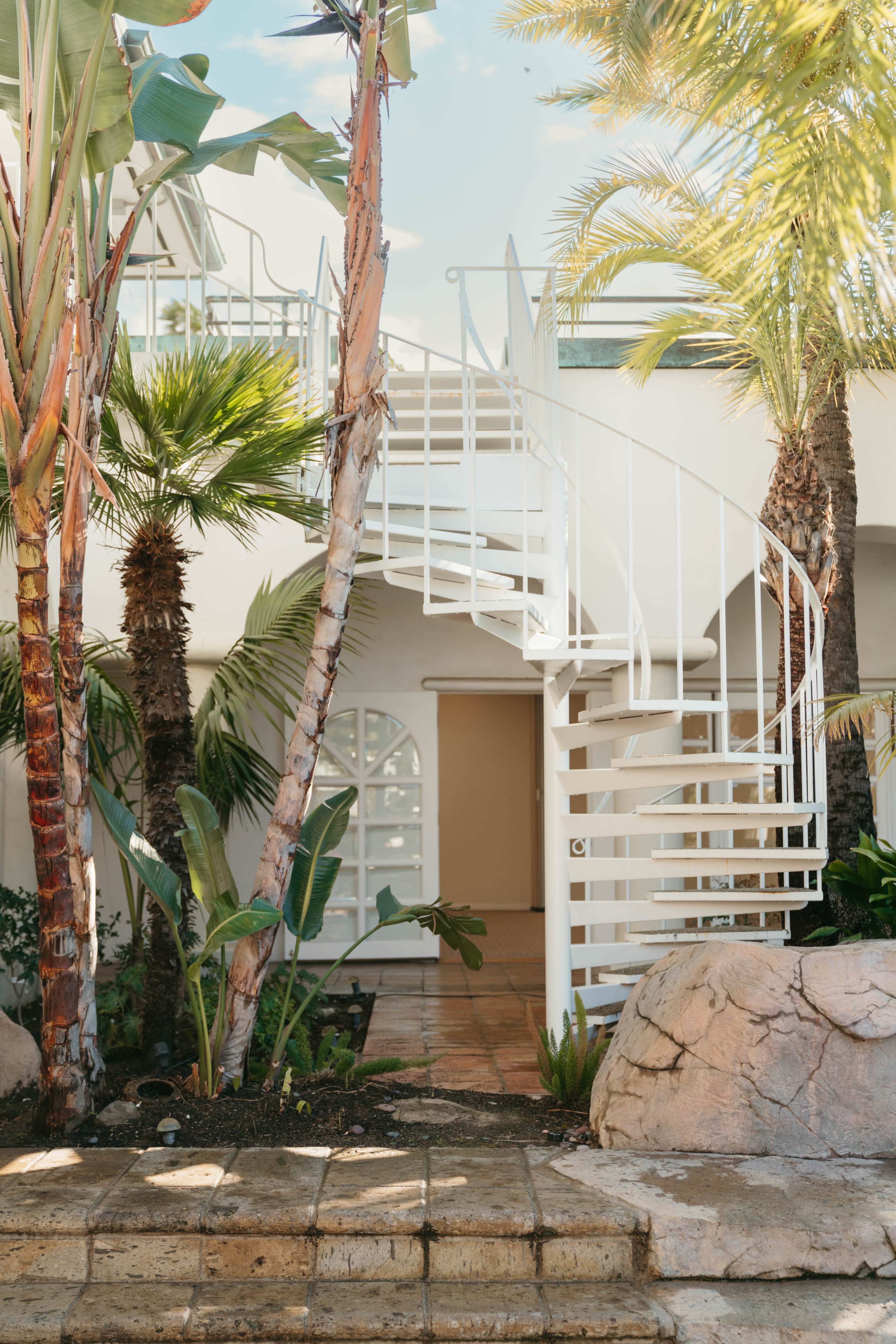 A white spiral staircase winds up beside palm trees and stone landscaping, leading to a building with a curved entrance.