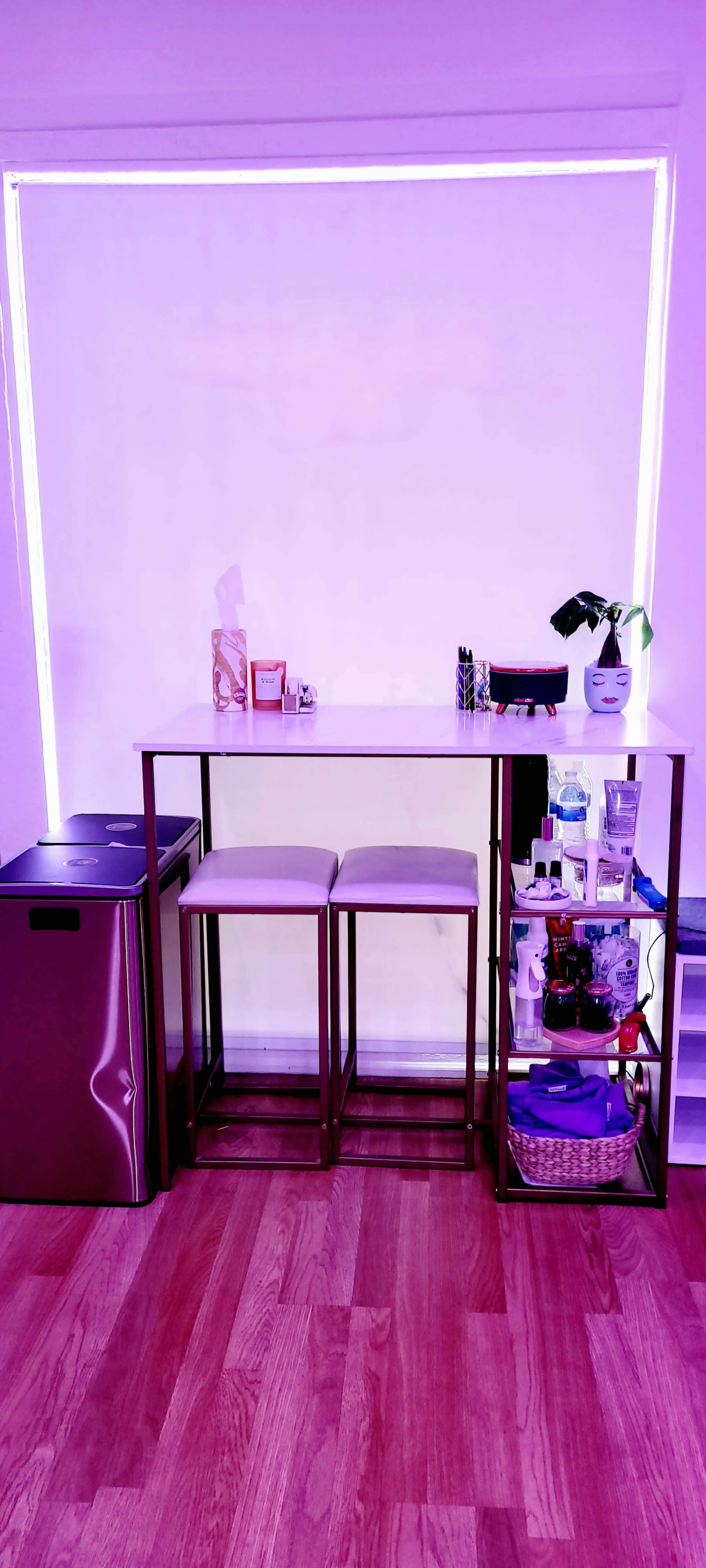 A small seating area features a high table with two stools, adjacent to a mini fridge, and a shelf displaying various items, all set against a softly lit background.