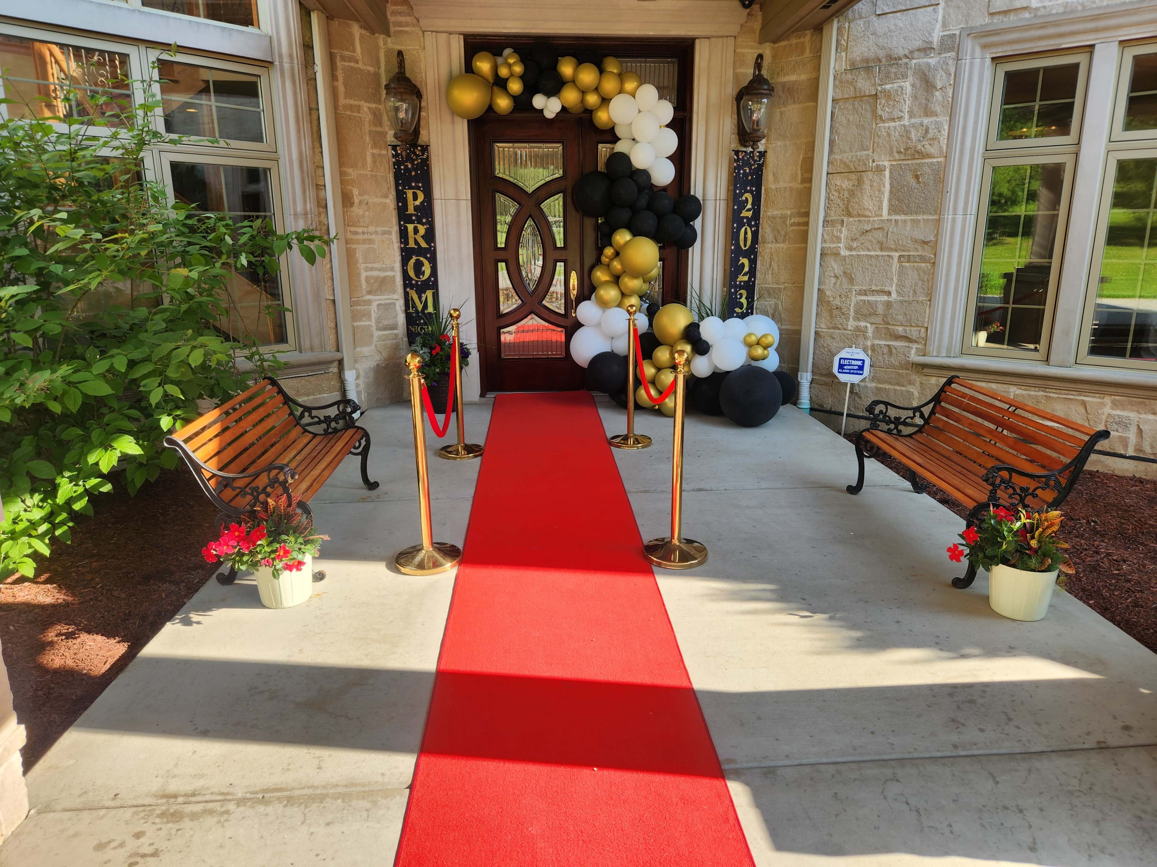 The entrance features a red carpet leading to ornate doors, flanked by benches and decorated with black, white, and gold balloons for a prom event.